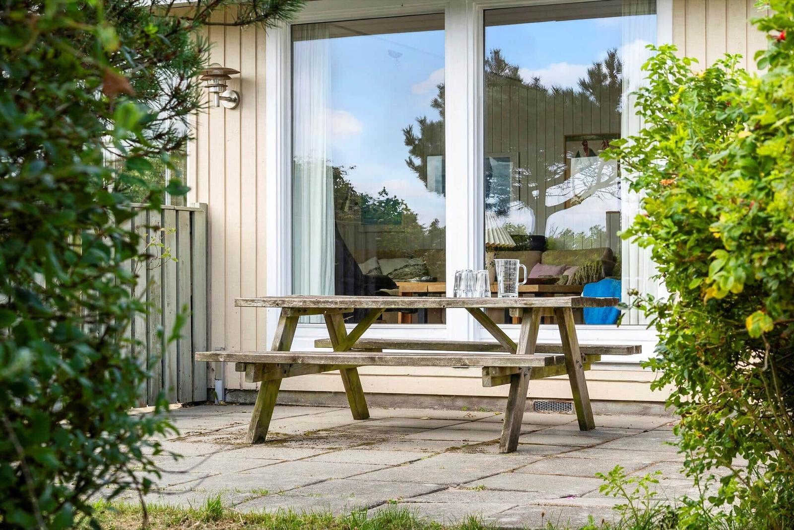 Holzpicknicktisch vor einem Haus mit großen Fenstern und grünen Pflanzen.
