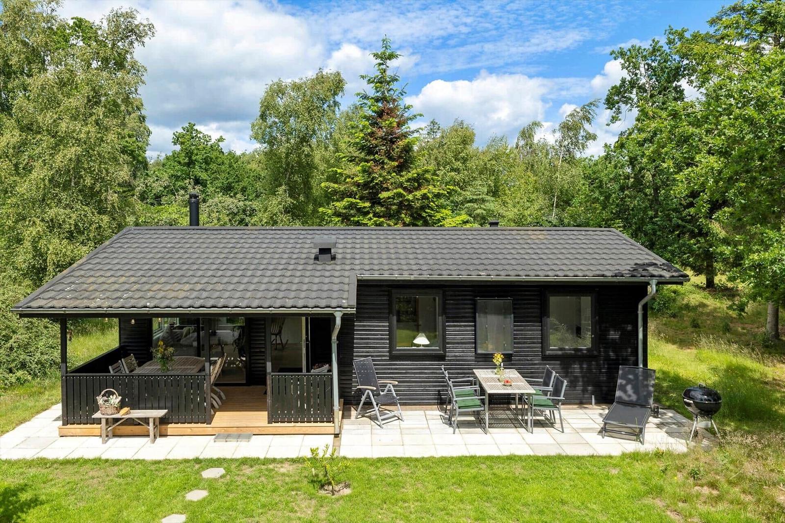 Holzhaus mit Terrasse, Grill und Garten. Umgeben von Bäumen unter blauem Himmel.