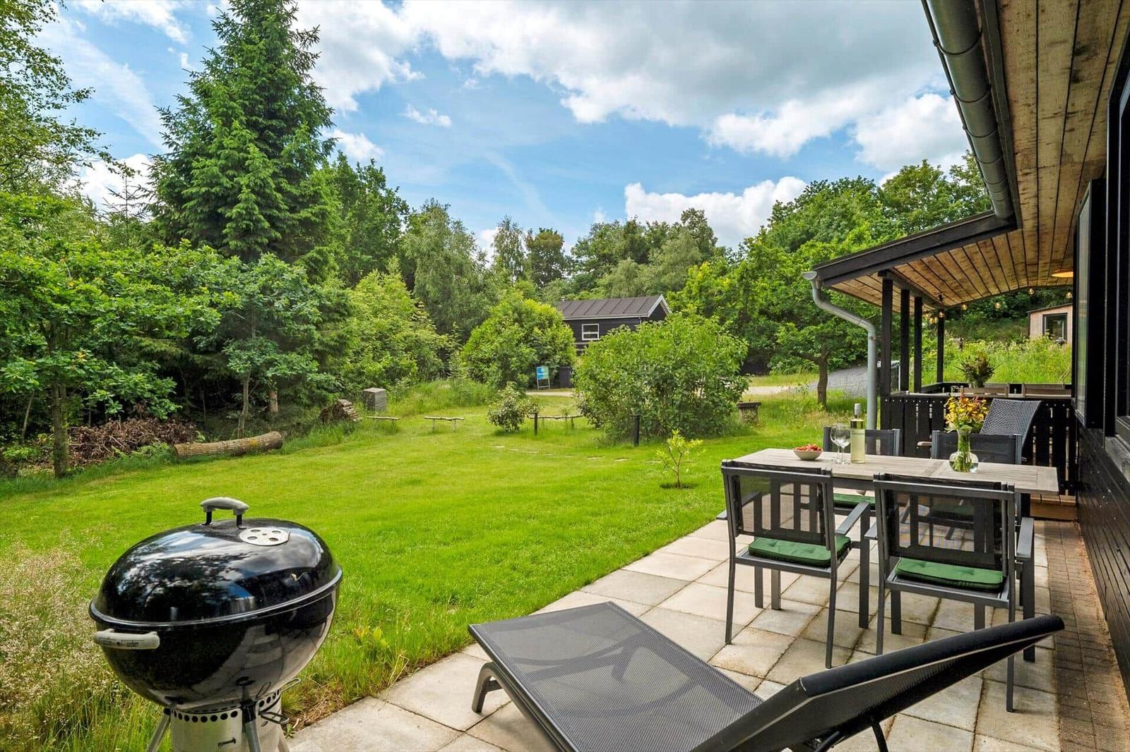 Terrasse mit Grill, Tisch und Stühlen. Grasfläche und Wald im Hintergrund.