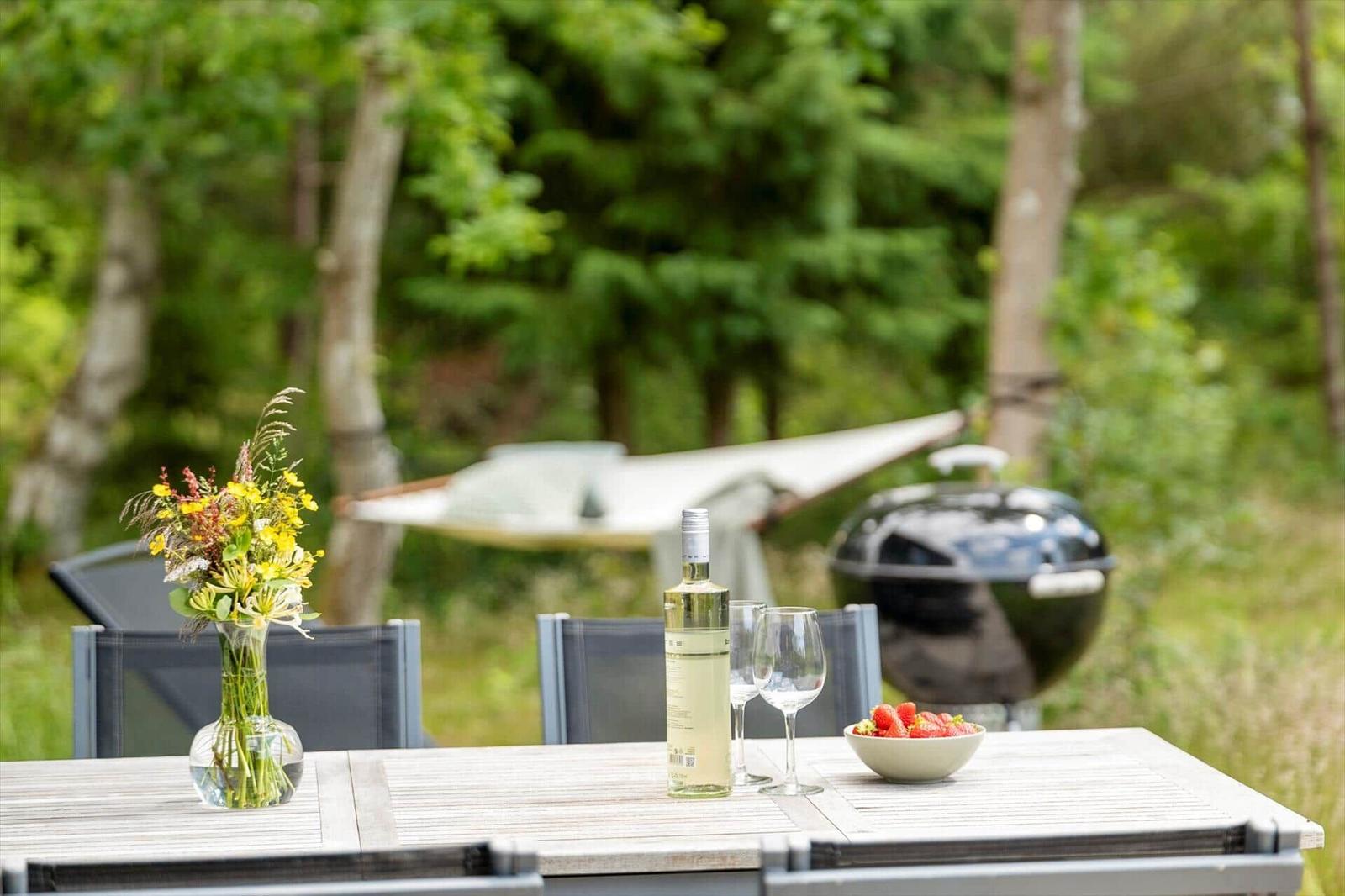 Holztisch mit Wein, Erdbeeren und Blumen im Garten. Grill und Hängematte im Hintergrund.