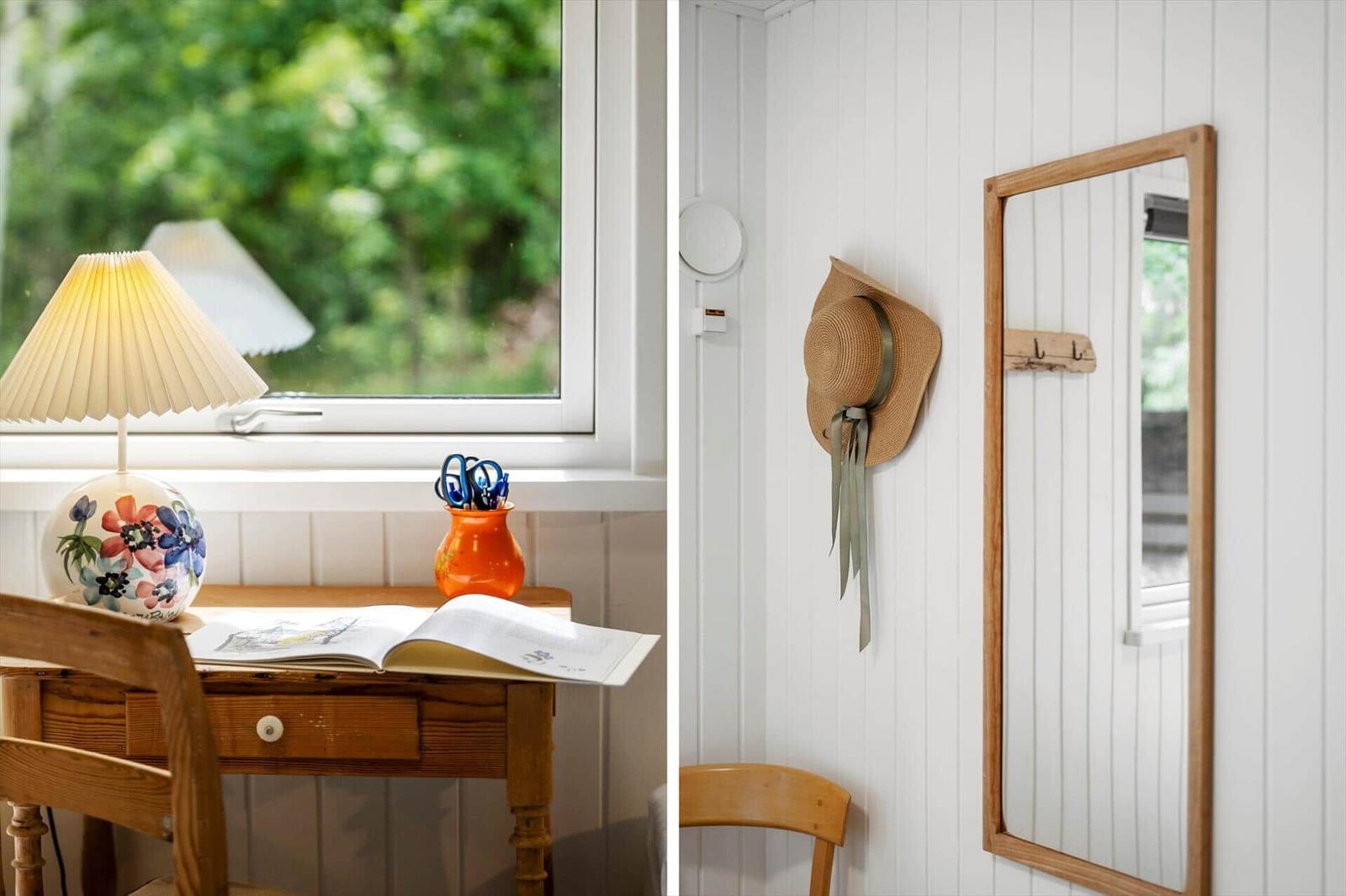 Schreibtisch mit Lampe und Buch neben Fenster. Spiegel und Hut an Wand.