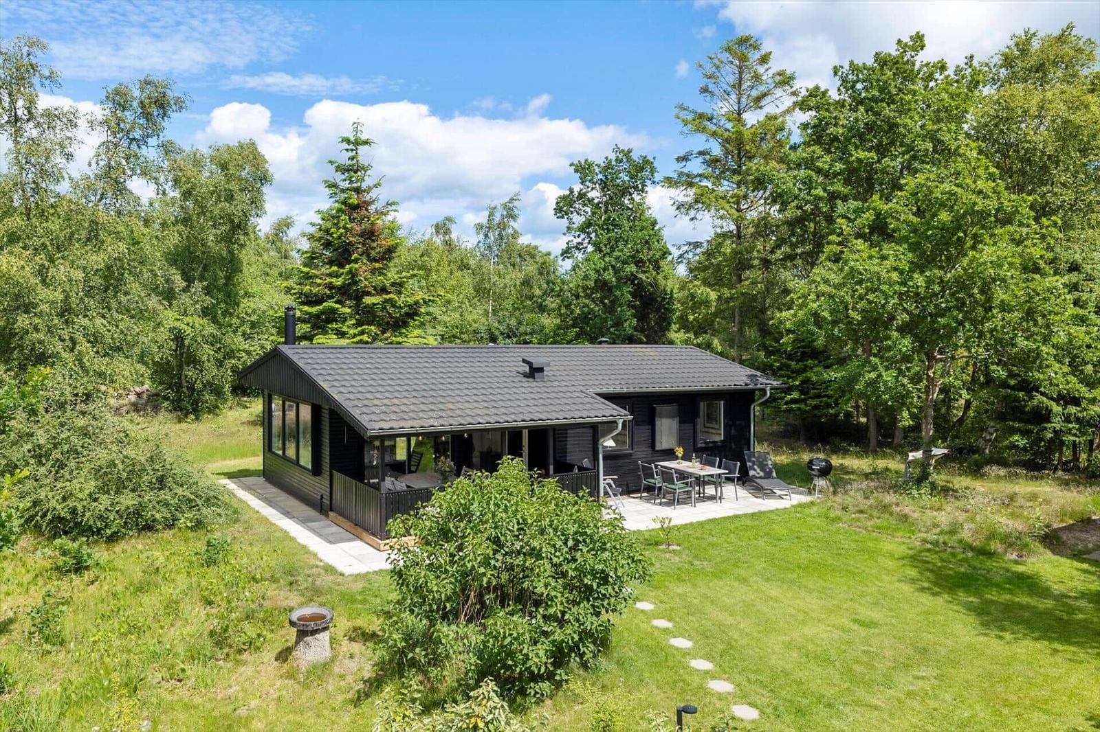 Schwarze Hütte mit Terrasse, Garten und Grillplatz zwischen Bäumen.