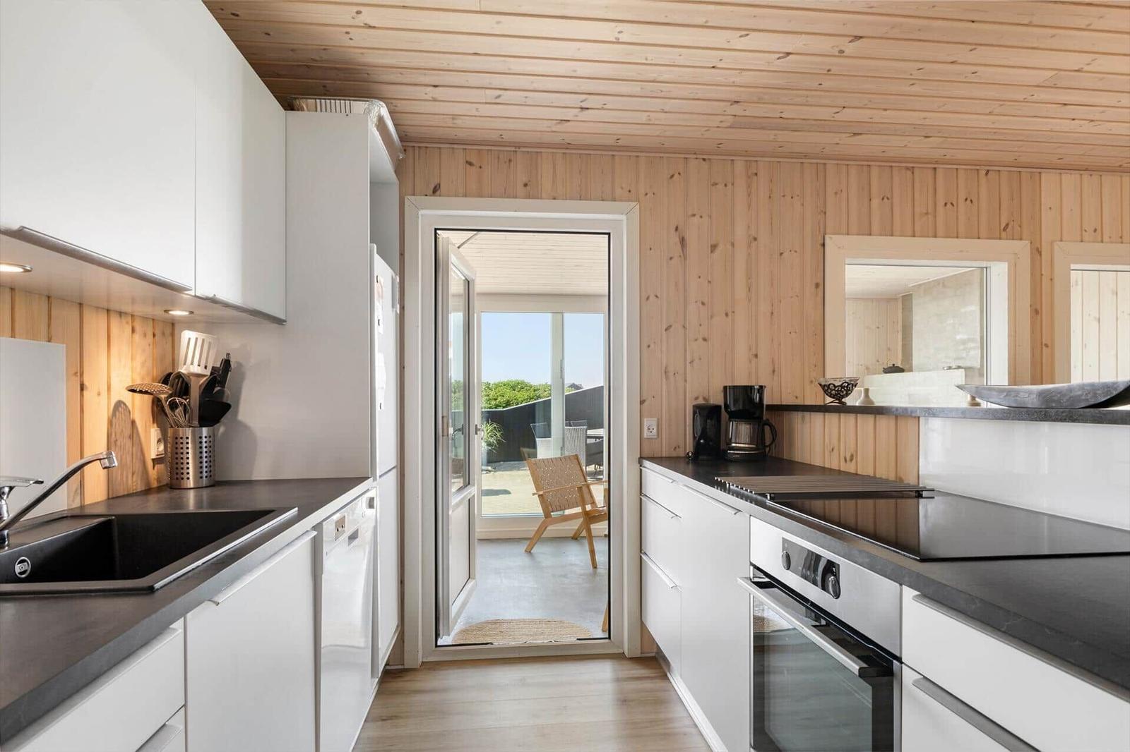 Küche mit weißen Schränken, dunklem Arbeitsplatz und Holzwand. Durchgang zur Terrasse mit Stuhl.
