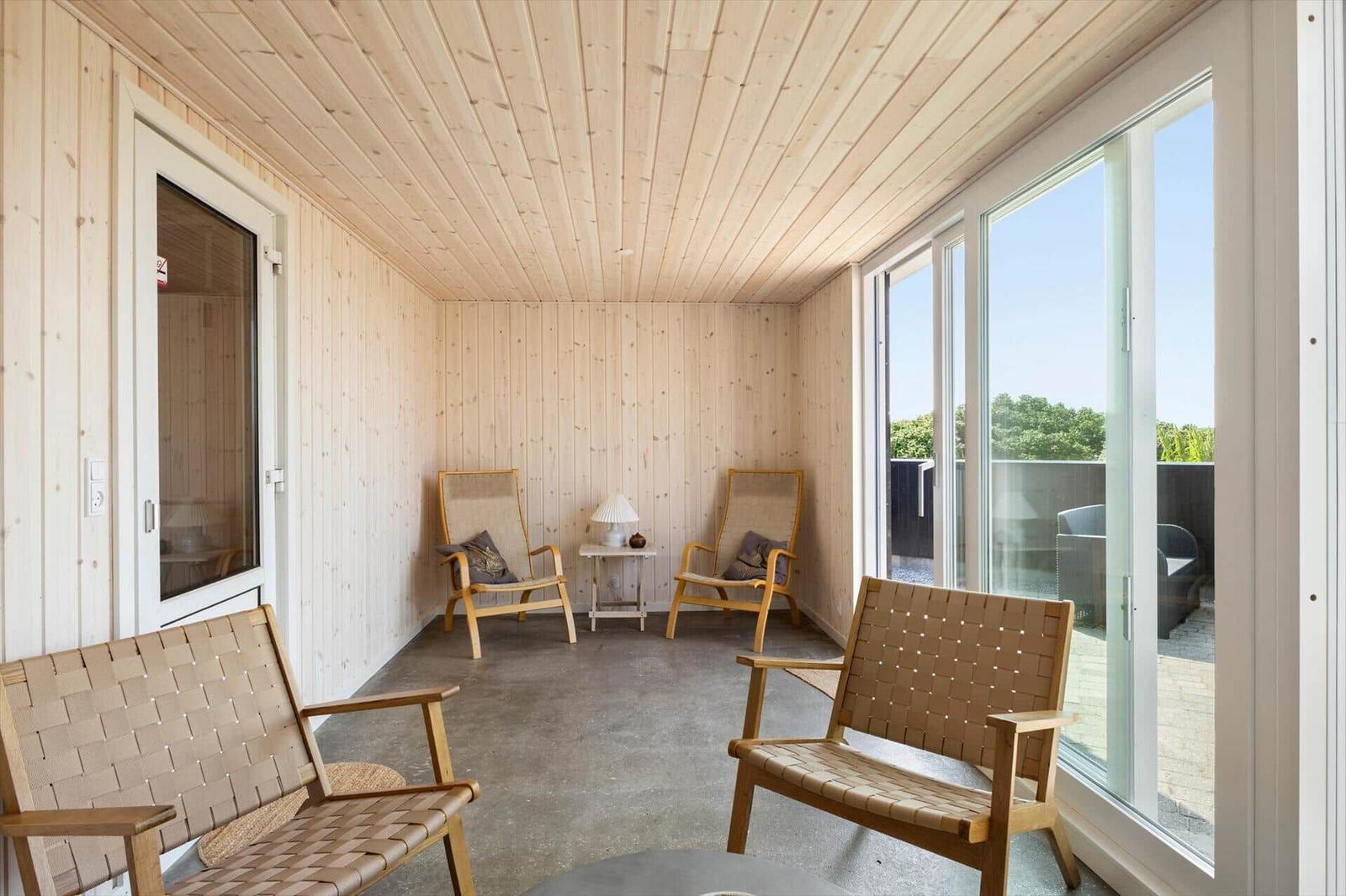 Ein Raum mit Holzwänden und -decke, zwei Sessel und einem Tisch. Große Fenster bieten Blick auf Außenbereich.