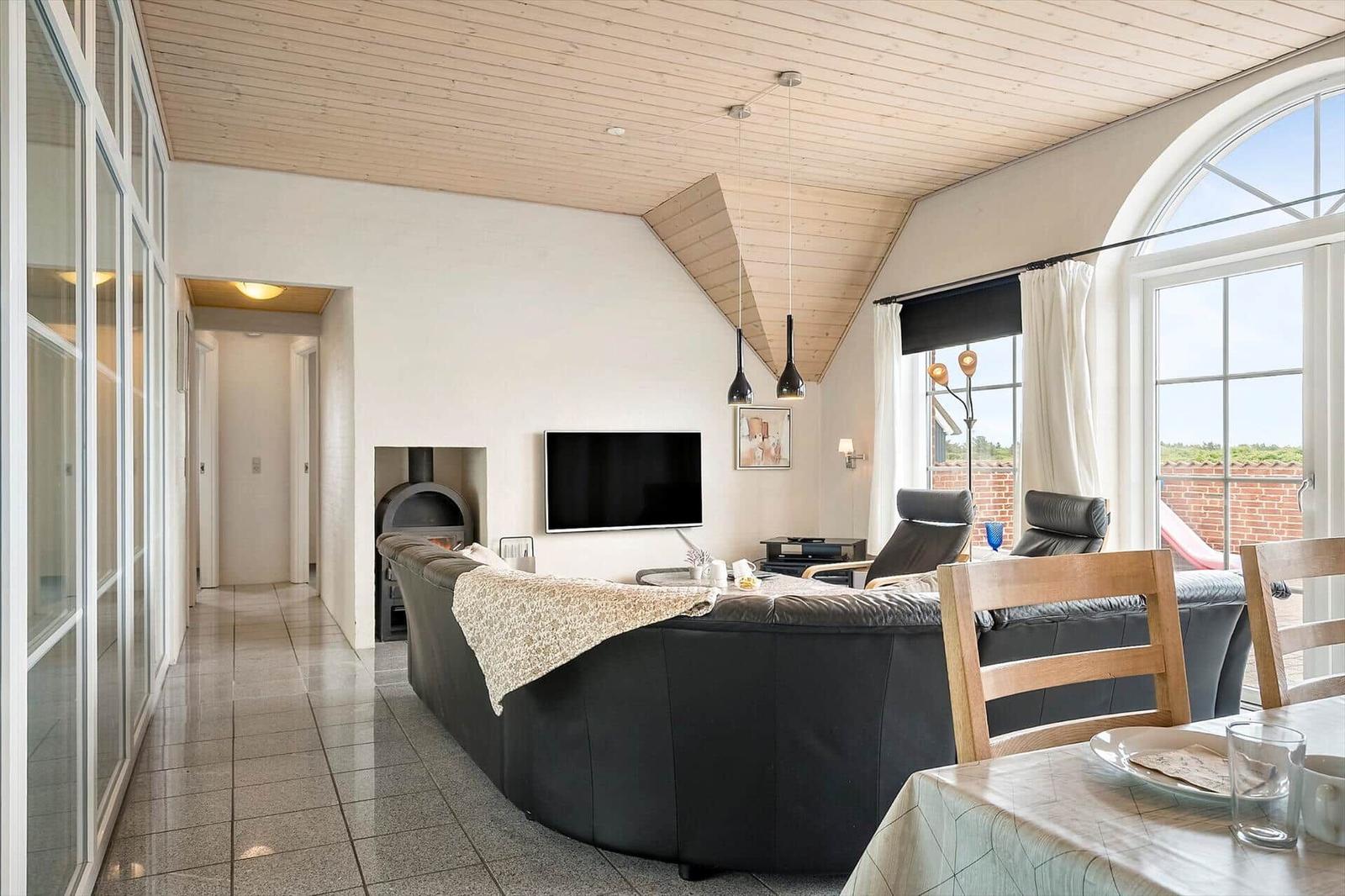 Wohnzimmer mit Sofa, Fernseher und Kamin. Große Fenster und Holzdecke.