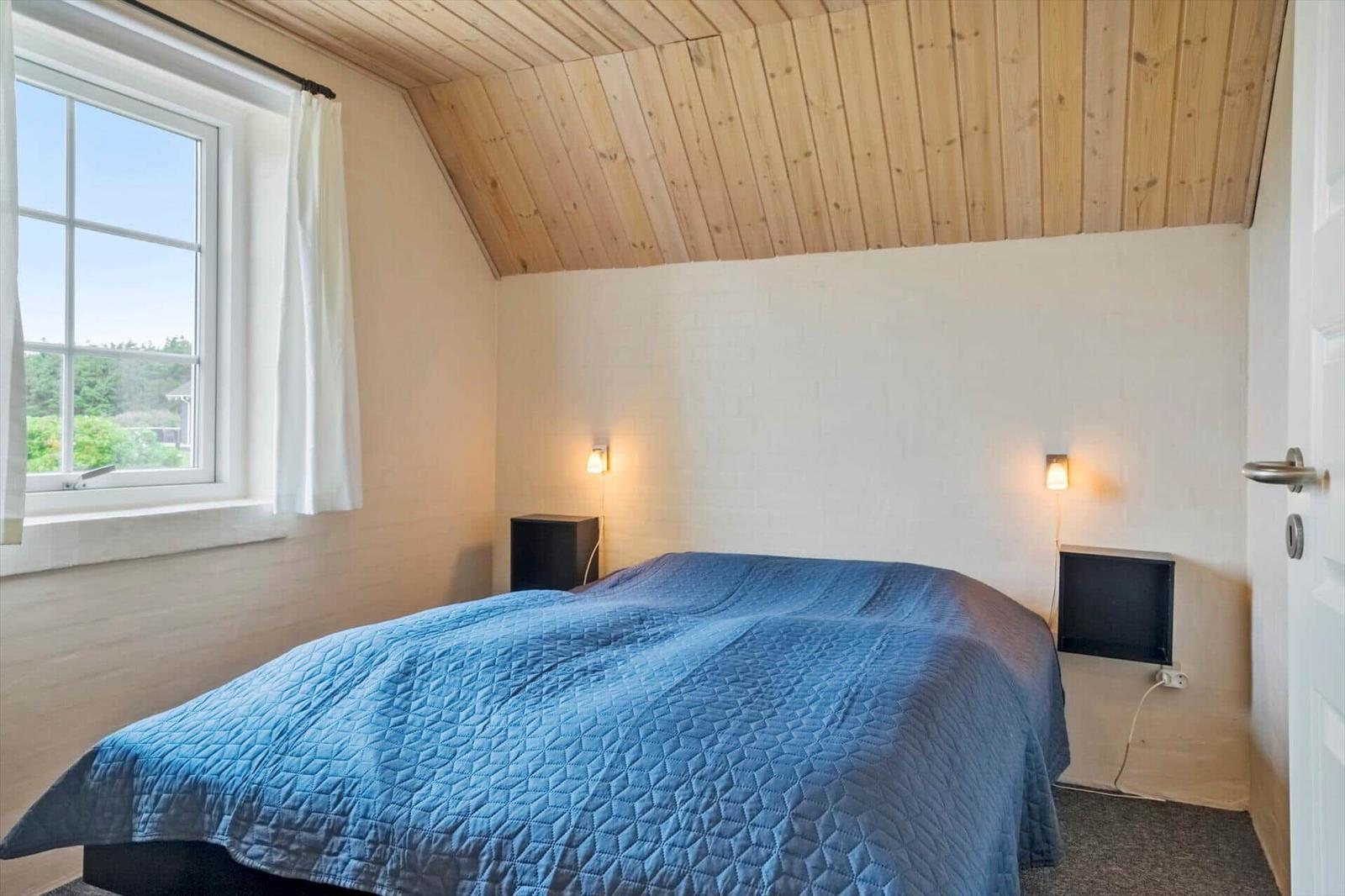 Schlafzimmer mit blauer Bettdecke, Holzdecke und Fenster mit Blick nach draußen.