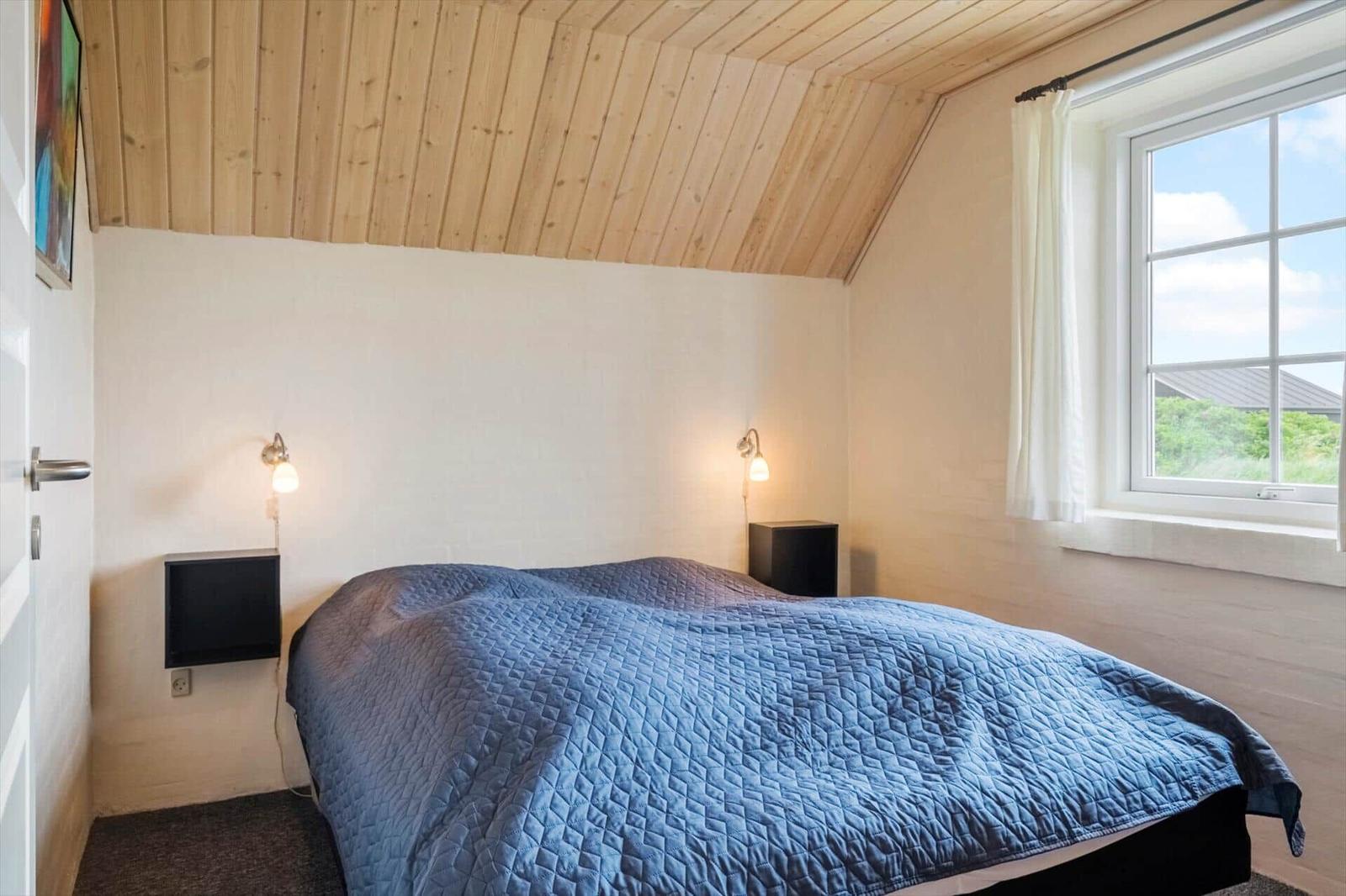 Schlafzimmer mit blauem Bett, Holzdecke und Fenster mit Blick nach draußen.
