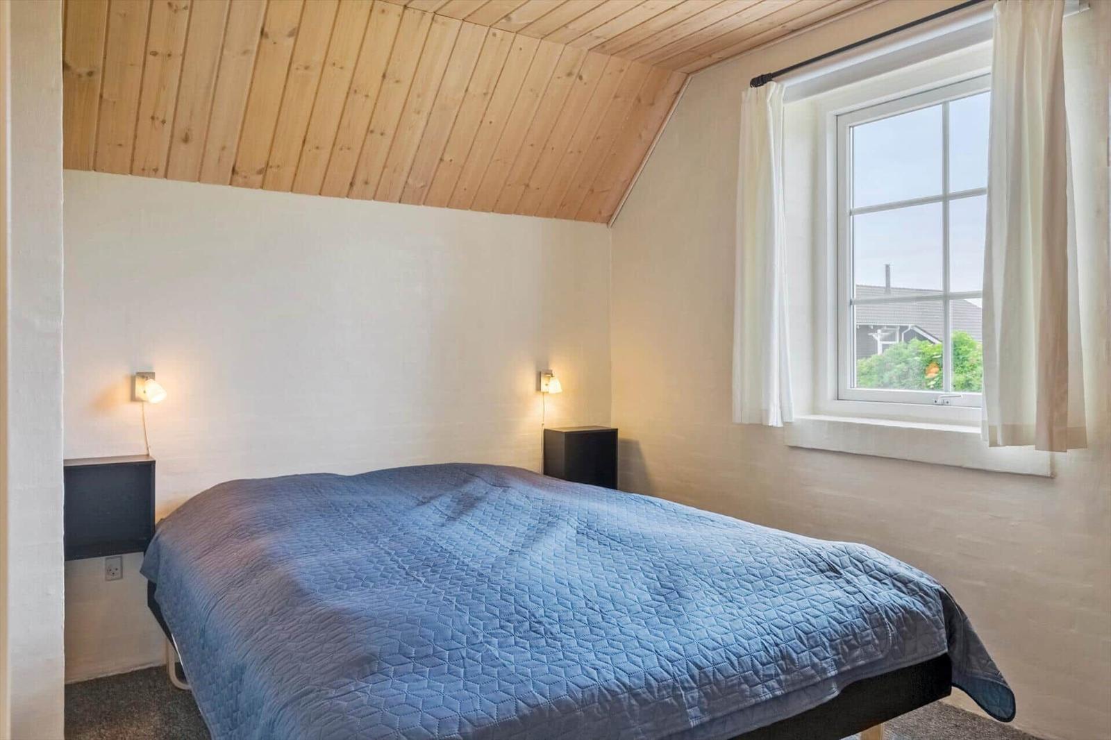 Schlafzimmer mit Bett, Holzdecke und Fenster. Zwei Wandlampen und Vorhänge.