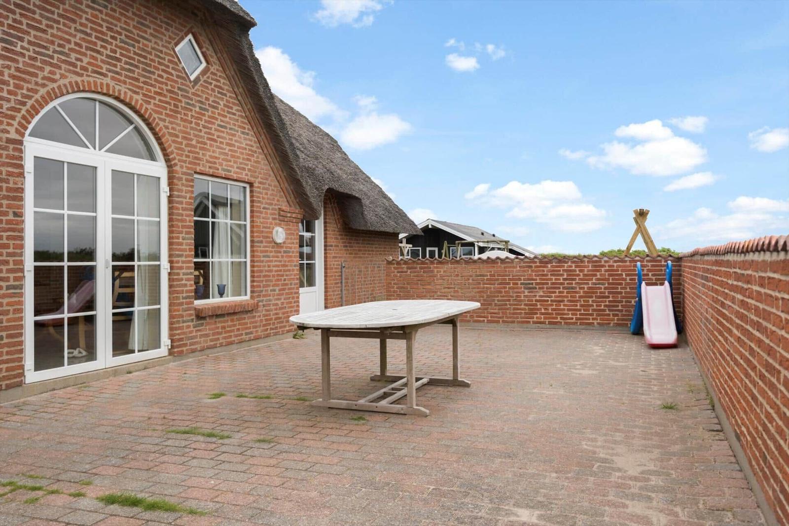 Terrasse mit Holztisch, Spielgeräten und Backsteinmauer vor einem Haus mit Dach aus Stroh.