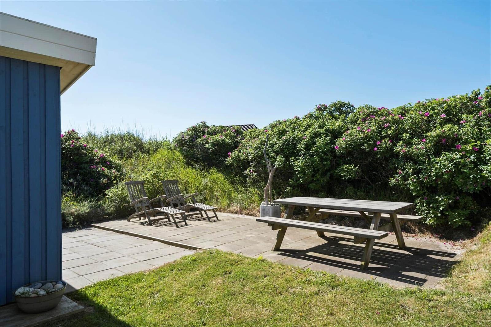 Terrasse mit Picknicktisch, zwei Stühlen und blauem Gebäude. Hintergrund mit grünen Sträuchern und rosa Blüten.