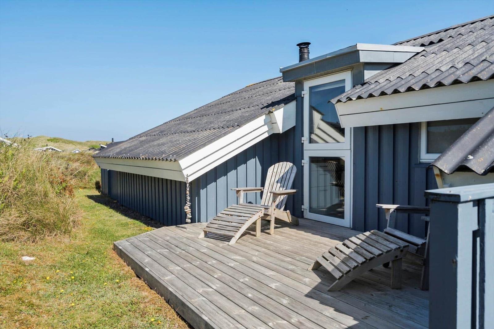 Holzterrasse mit Liegestühlen vor einem grauen Strandhaus mit Dach und Fenstern.