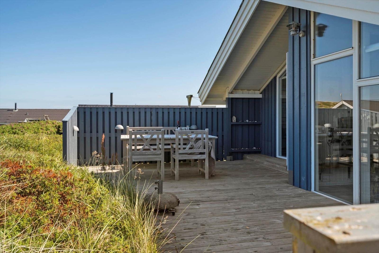 Terrasse mit Holztisch und Stühlen neben blauem Haus. Grasland im Hintergrund.