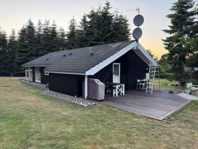 Black wooden house with terrace, grill, and satellite dishes. Surrounded by trees and grass.