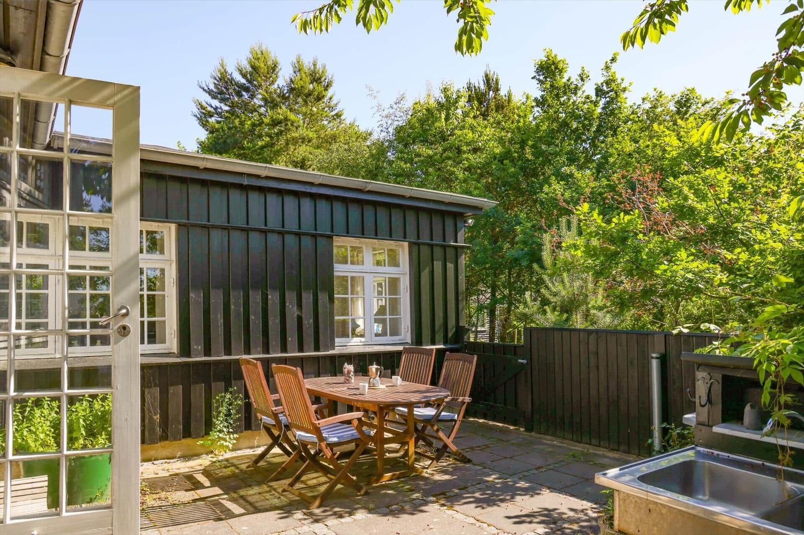 Backyard with wooden table, chairs, and grill. Dark wood structure and green trees in the background.
