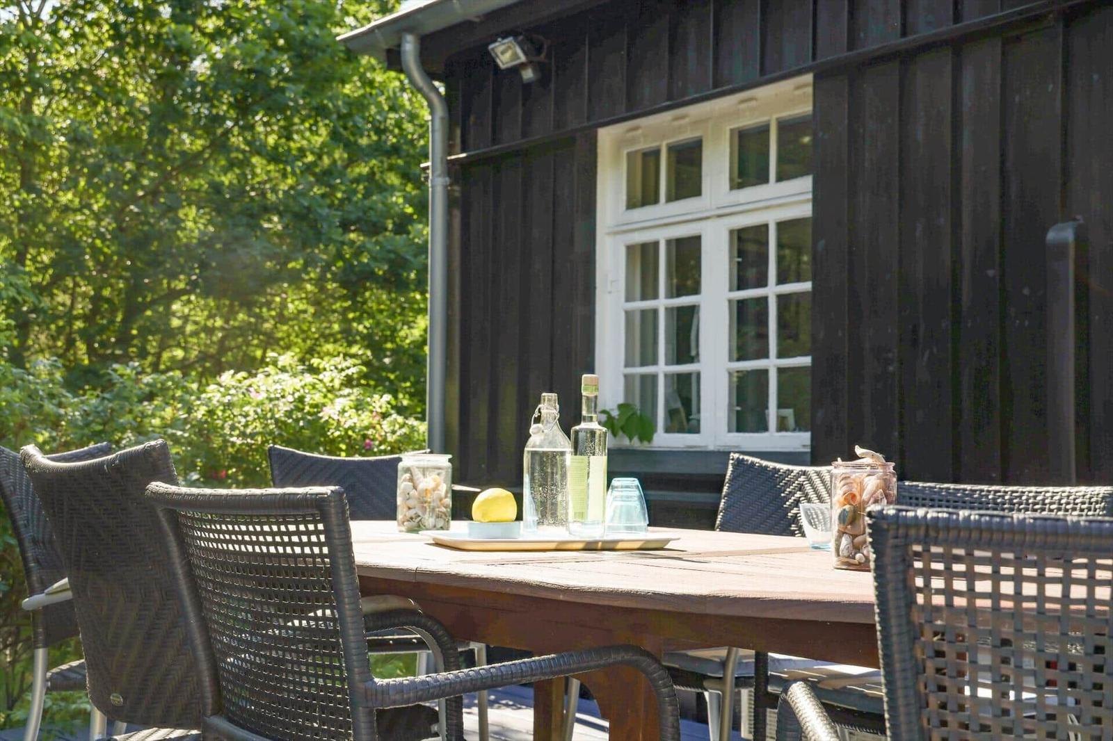 Wooden table with chairs on terrace in front of dark wooden house with white window.