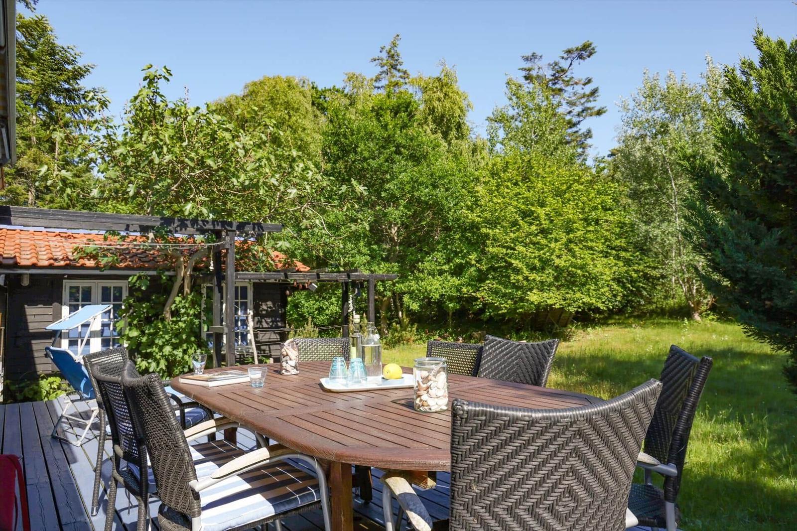 Terrace with table and chairs, surrounded by greenery and a house in the background.