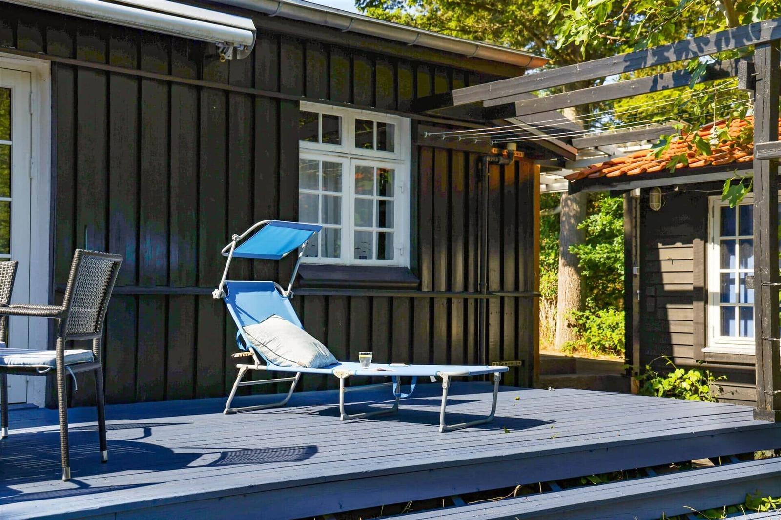 Deck with blue lounge chair, chair, and glass. Wooden house with white windows.
