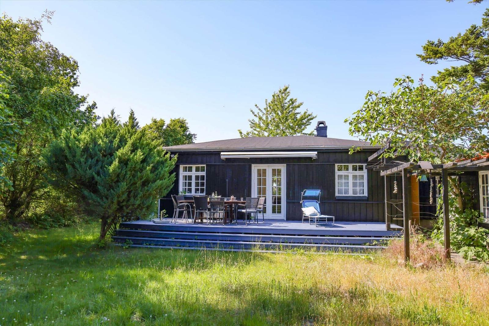 A dark wooden house with a terrace, garden, and trees. There is a table with chairs and a lounge chair.