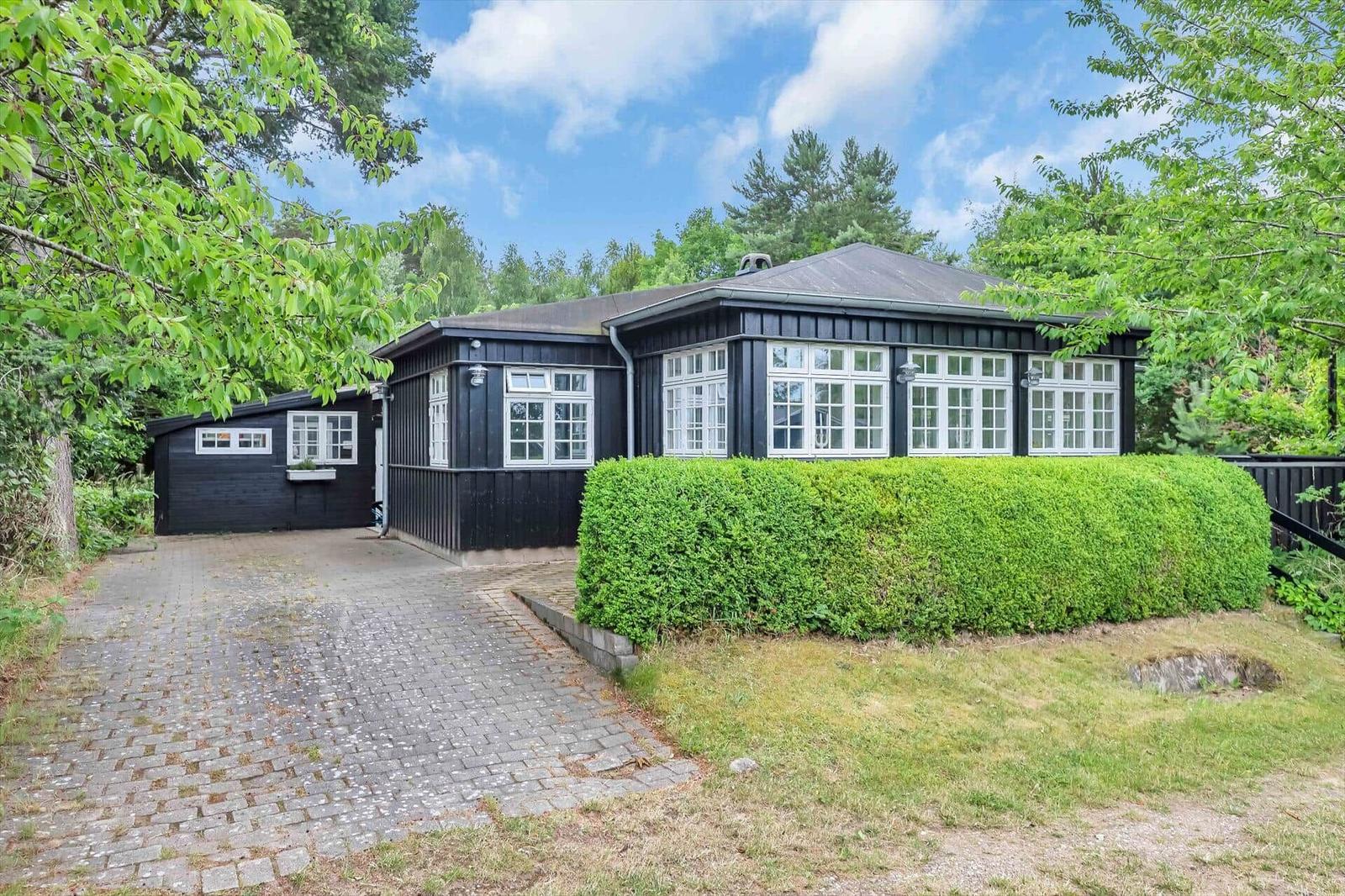Black wooden house with white windows and green hedge. Stone path and trees in the background.