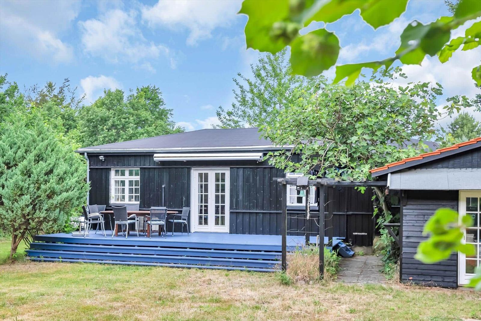 Black wooden house with terrace and garden. White windows and door. Surrounded by trees.