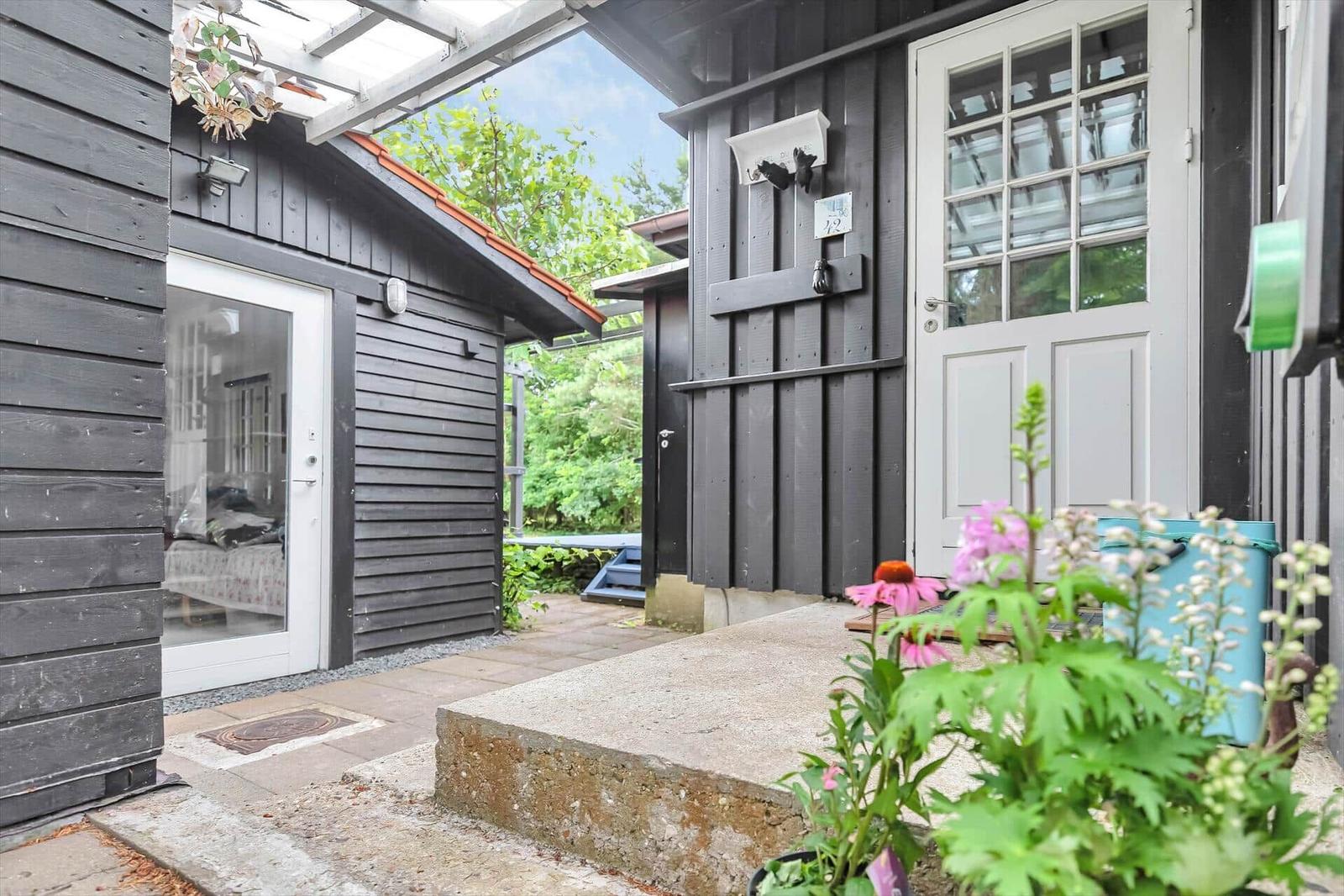 Entrance with gray wooden facade, white door, and flower beds.