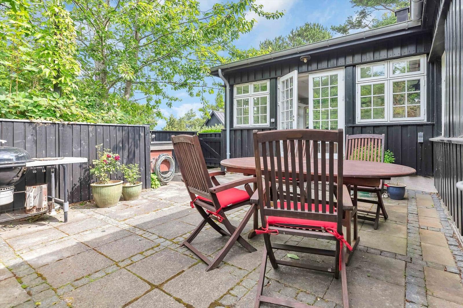Backyard with table, chairs, and grill. Black wooden wall and windows visible.