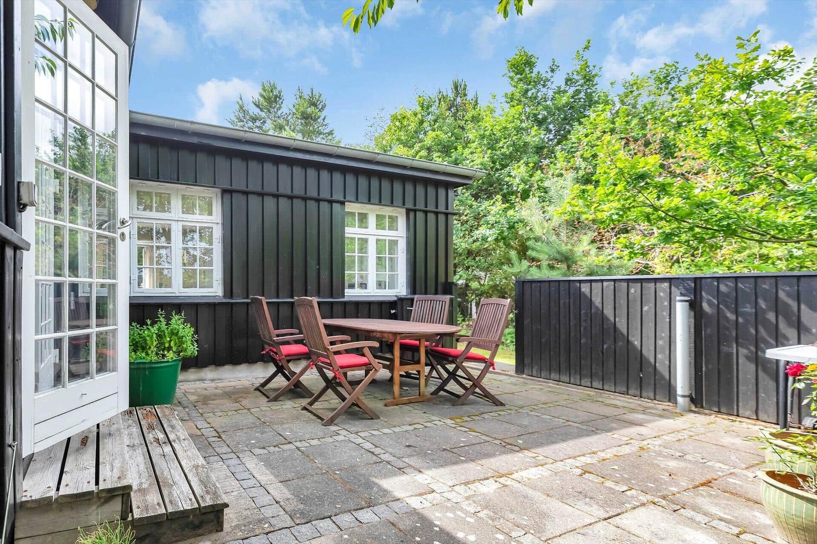 Terrace with table, chairs, and plants. Backyard with black wood siding and trees.