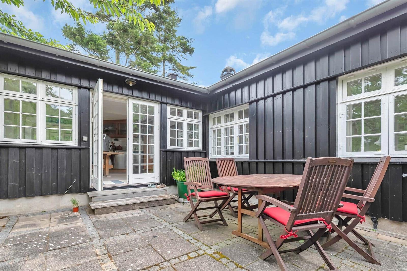 Terrace with wooden table and chairs, black siding, white windows, open door to interior.