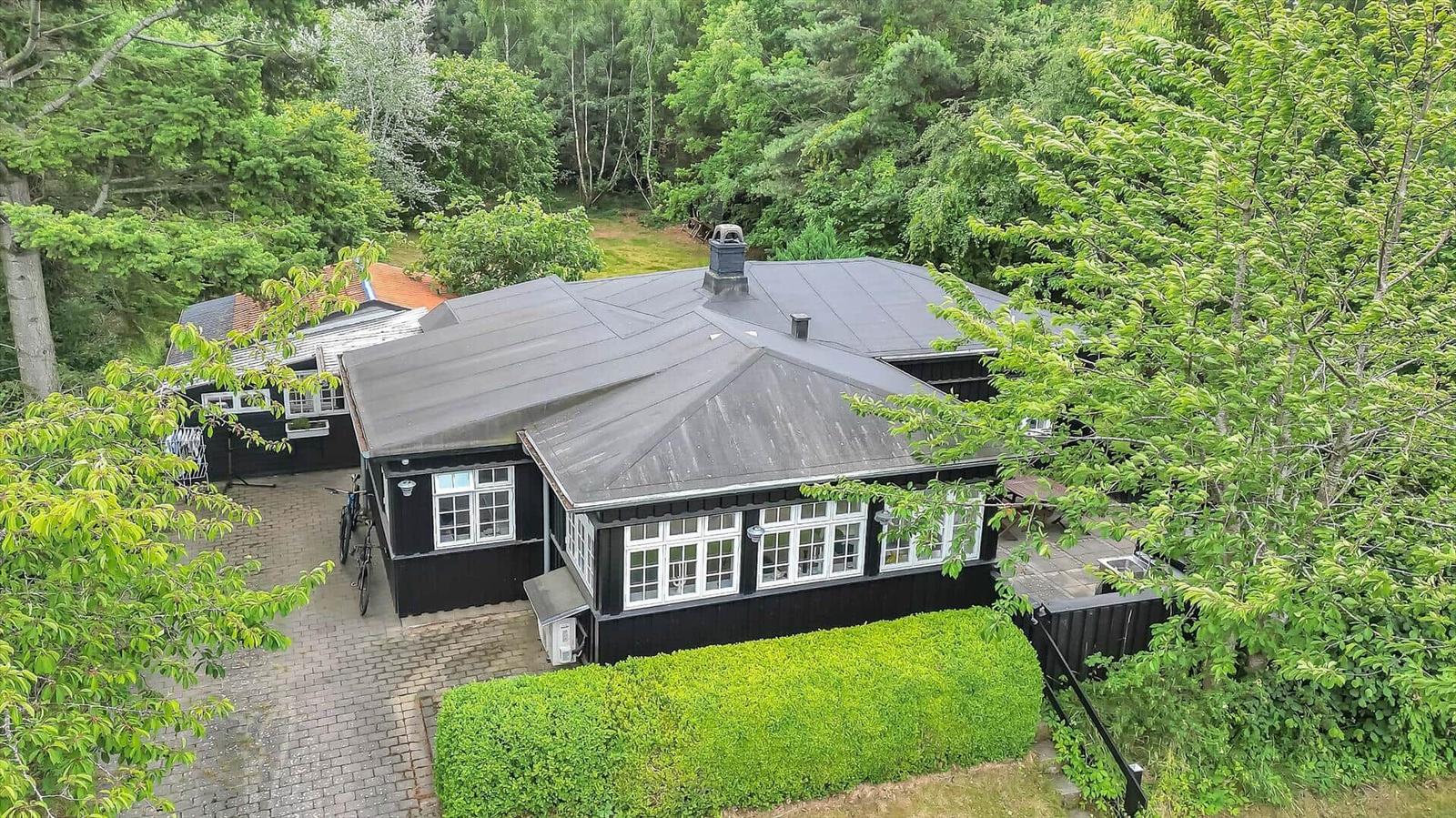 Black wooden house with gray roof, surrounded by green trees and a paved yard.