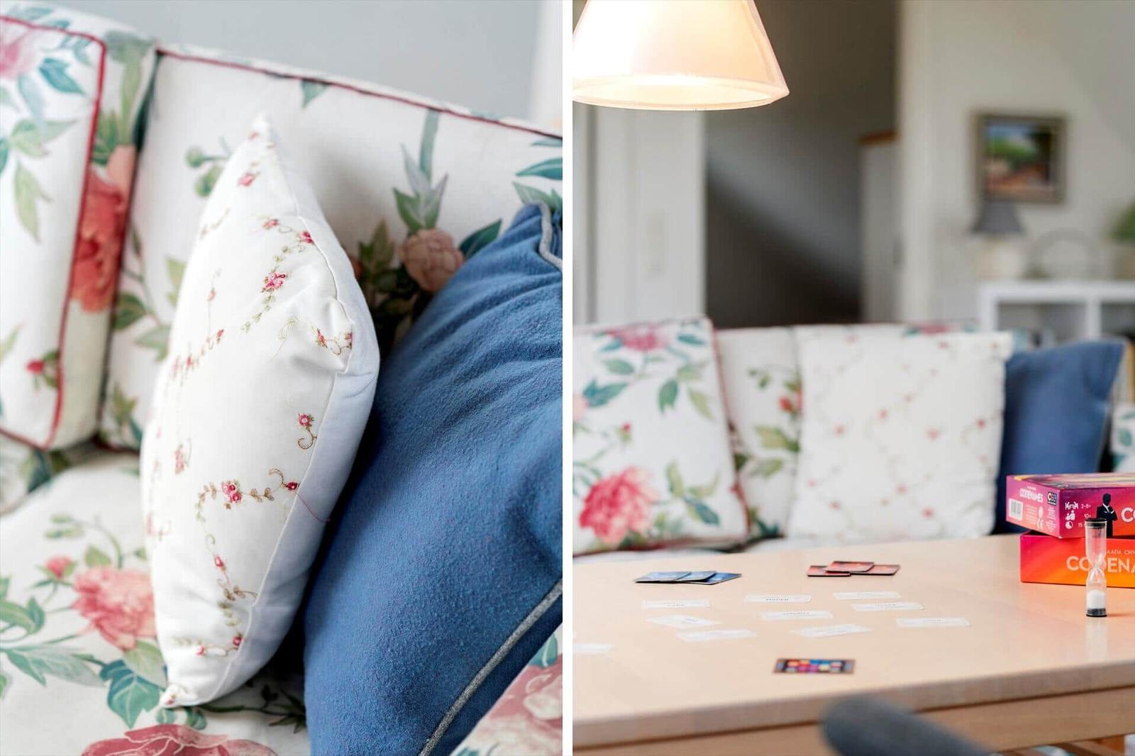 Sofa mit floralen Kissen und blauem Kissen. Tisch mit Spielkarten und Spielbox.