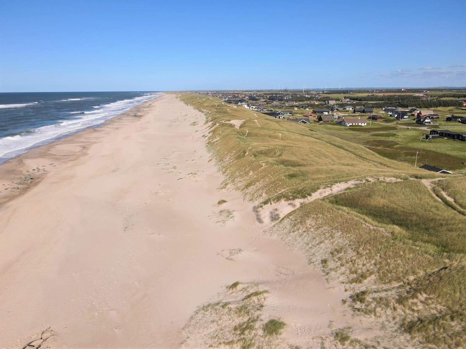 Strand mit Dünen und Siedlung im Hintergrund unter blauem Himmel.
