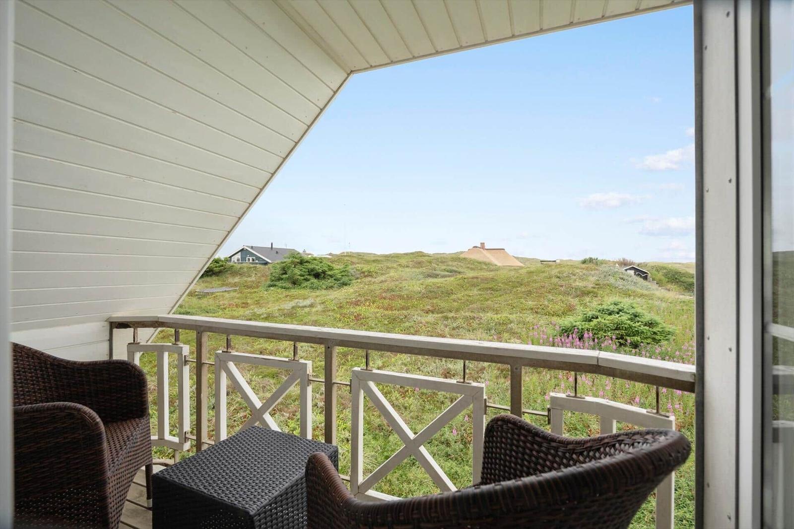 Balkon mit zwei Stühlen und Tisch, Blick auf grüne Dünen und Himmel.