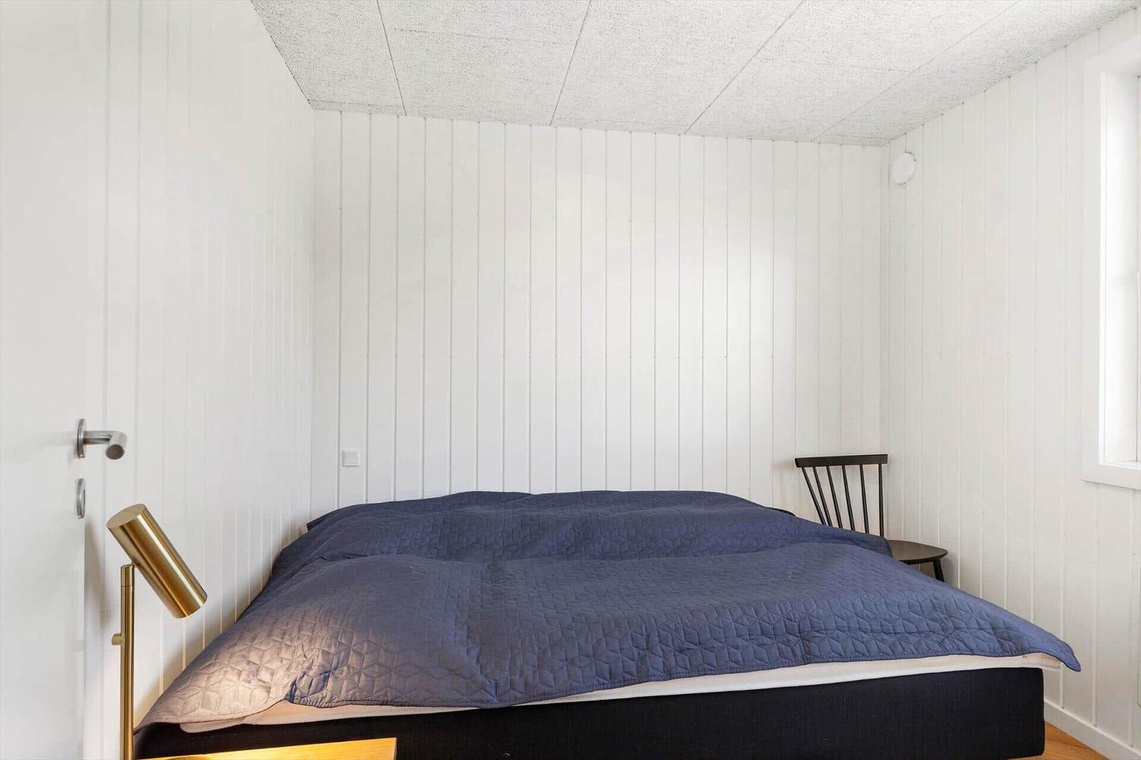 Schlafzimmer mit Bett, Nachttisch und Stuhl. Wände sind weiß verkleidet.