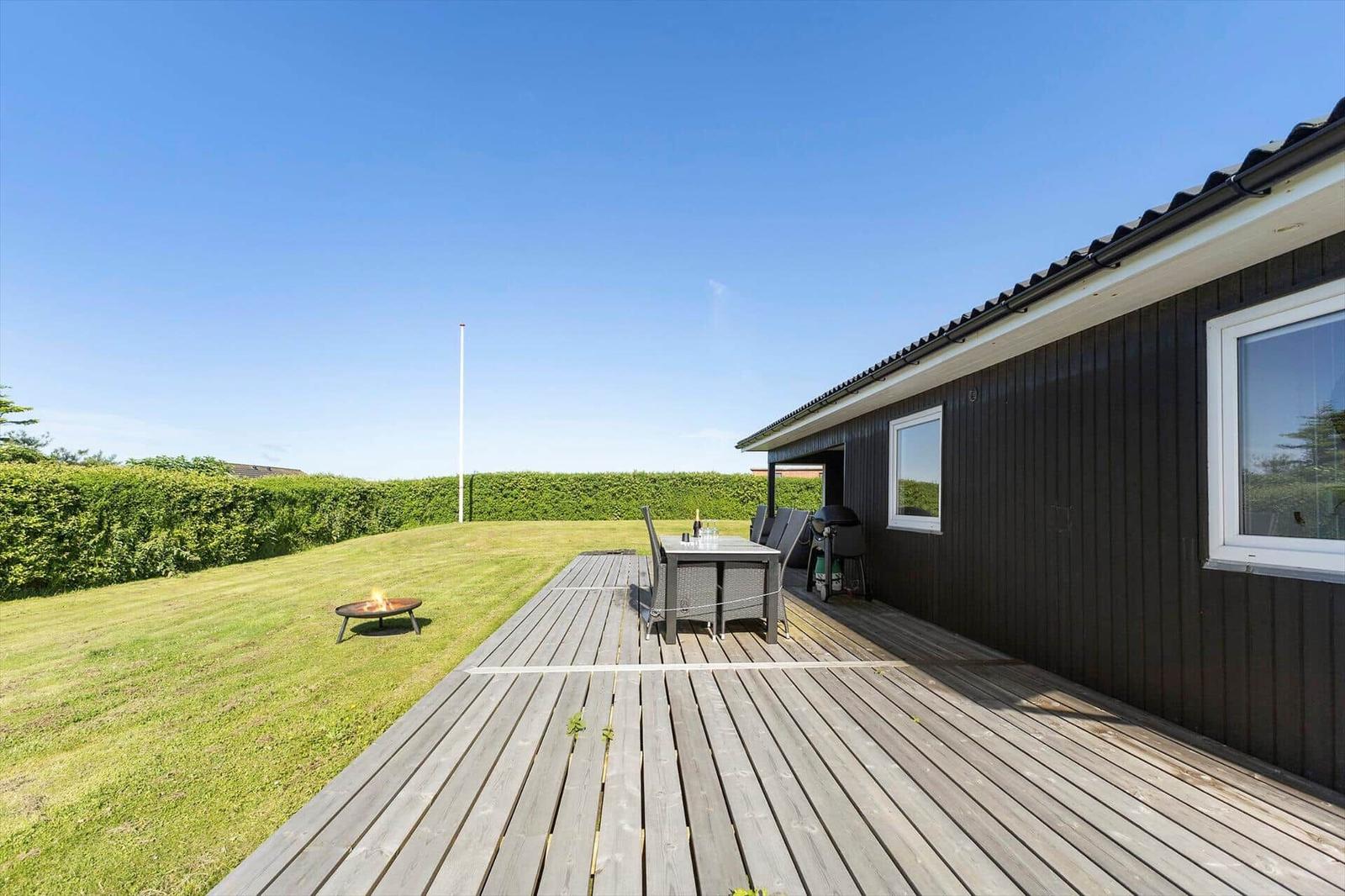 Terrasse mit Tisch, Stühlen und Grill neben dunkelbraunem Haus mit grünem Rasen und Heckenzaun.