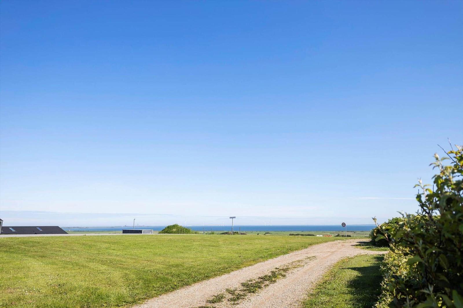 Grünes Feld mit unbefestigter Straße, Blick auf das Meer und Windräder unter blauem Himmel.
