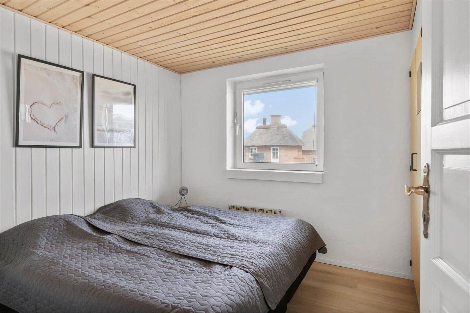 Bedroom with bed, window, and wooden ceiling. Wall art and door visible.