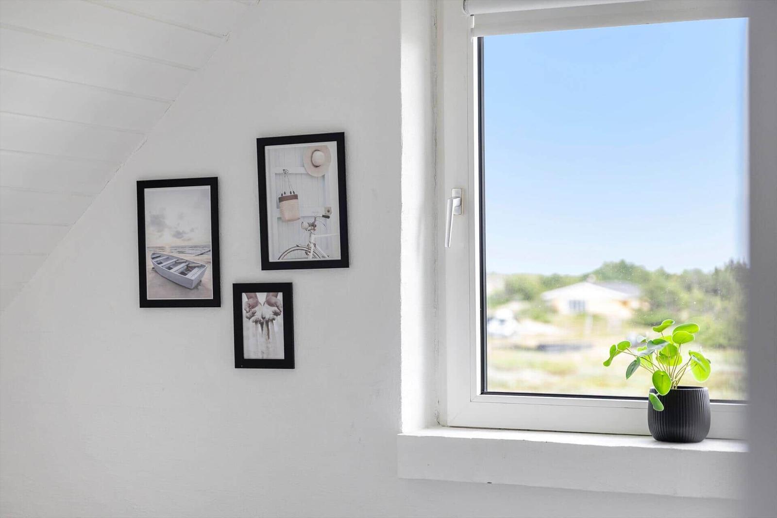 A window with a view of green areas. Three framed pictures on the wall. A plant on the windowsill.