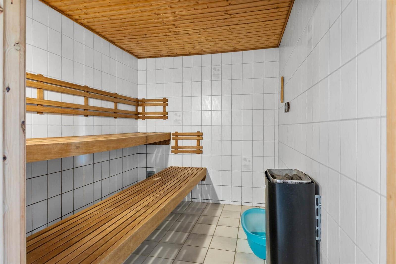 Sauna with wooden benches and white tiled walls. Heater and bucket are present.