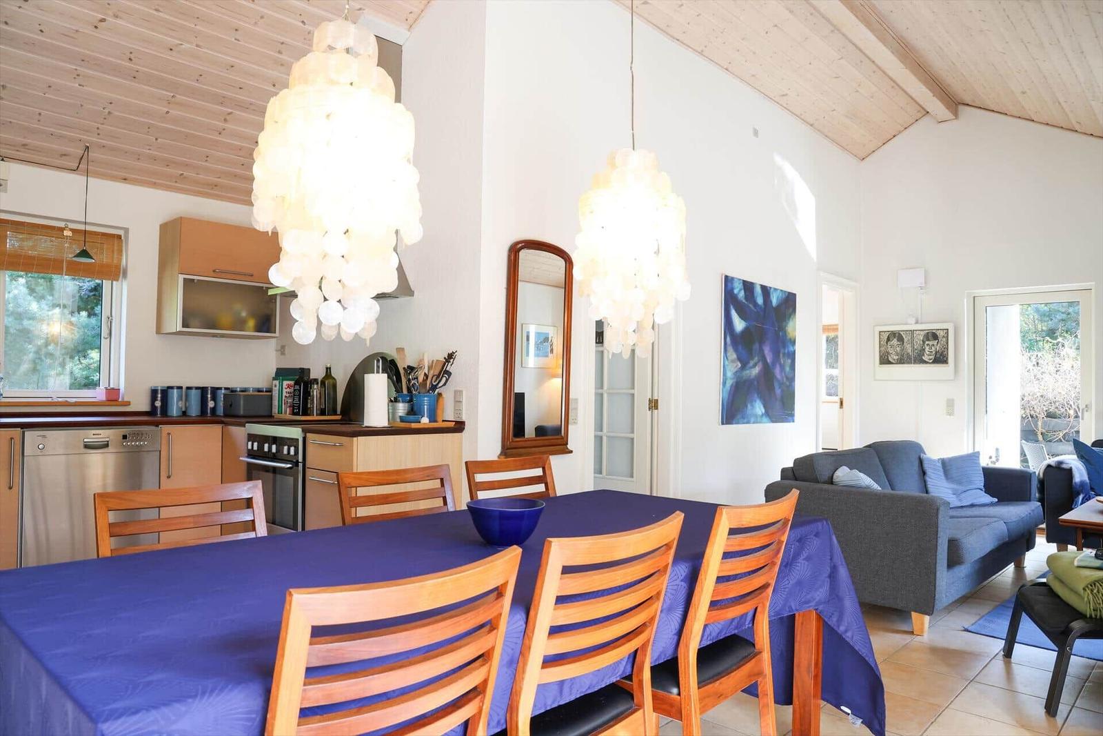 Essbereich mit blauer Tischdecke und Holzstühlen unter hängenden Lampen. Wohnbereich mit Sofa und Wandbild.