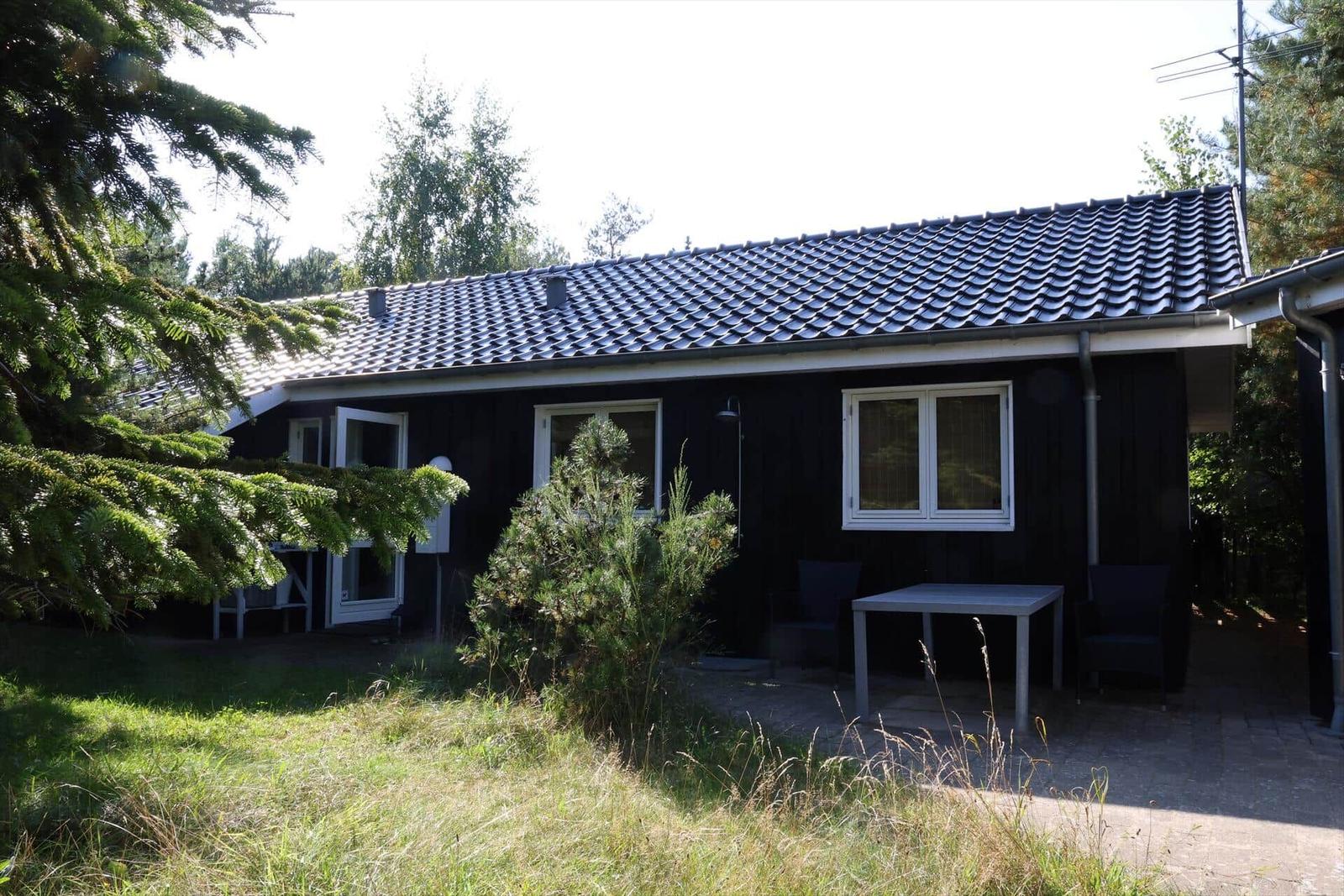 Schwarzes Holzhaus mit Terrasse, Tisch und Stühlen. Umgeben von Bäumen und Gras.