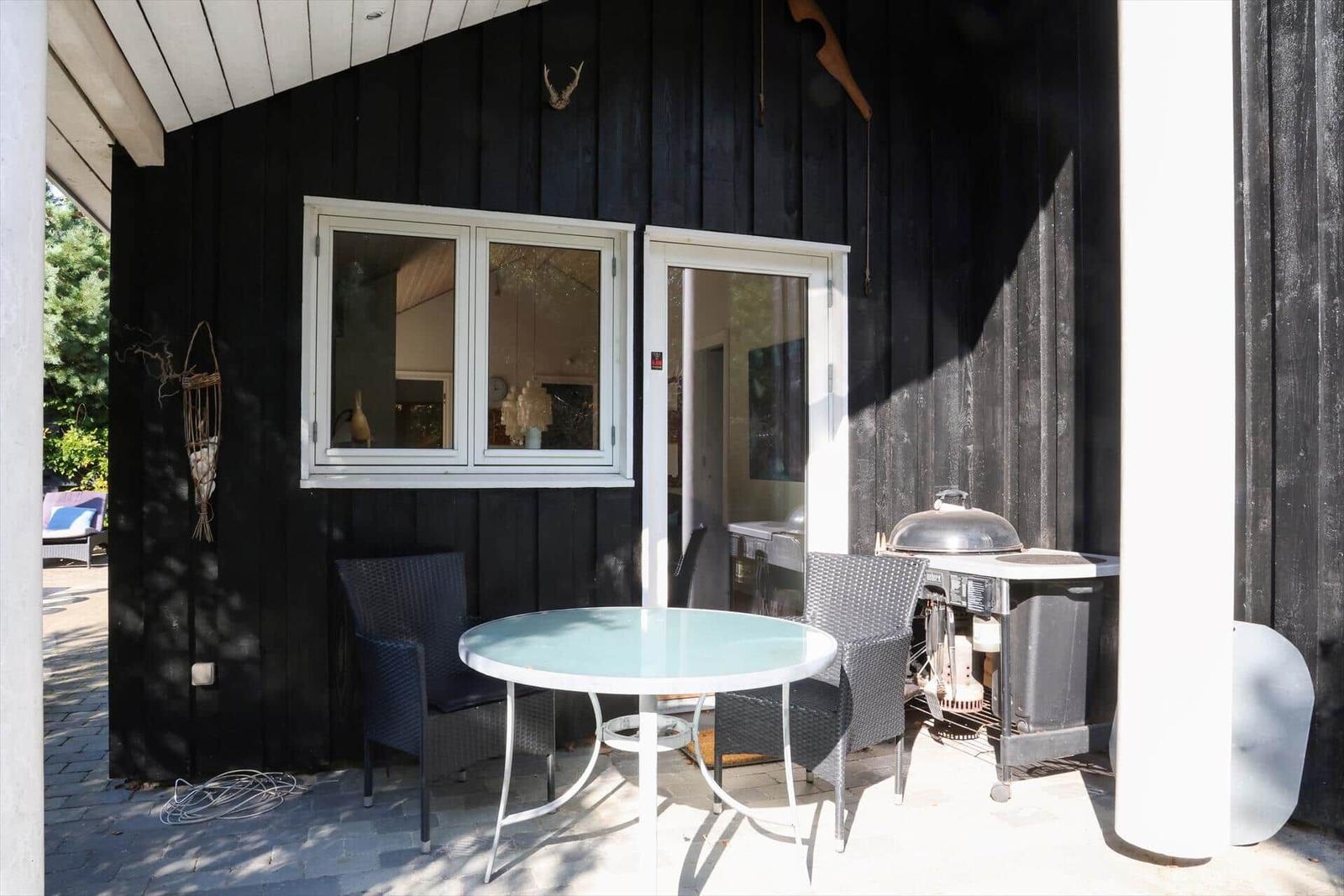 Außenbereich mit Tisch, Stühlen und Grill vor schwarzem Holzhaus mit Fenster und Tür.