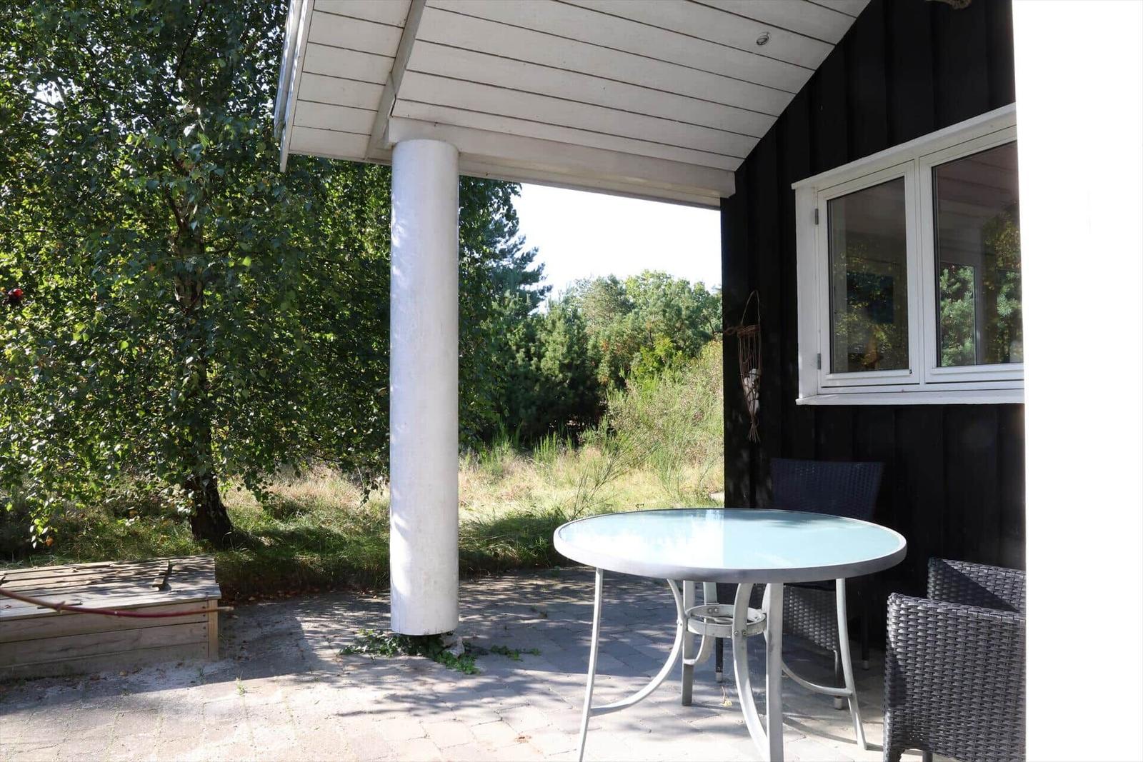 Terrasse mit Tisch und Stühlen unter Dach. Hintergrund: Wald und Gras.