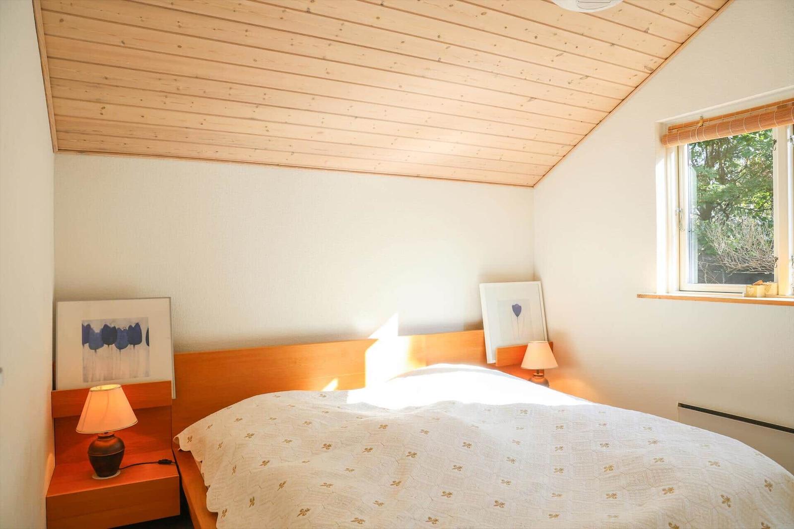 Schlafzimmer mit Holzdecke, Bett und Fenster. Auf Nachttischen stehen Lampen und Bilder.