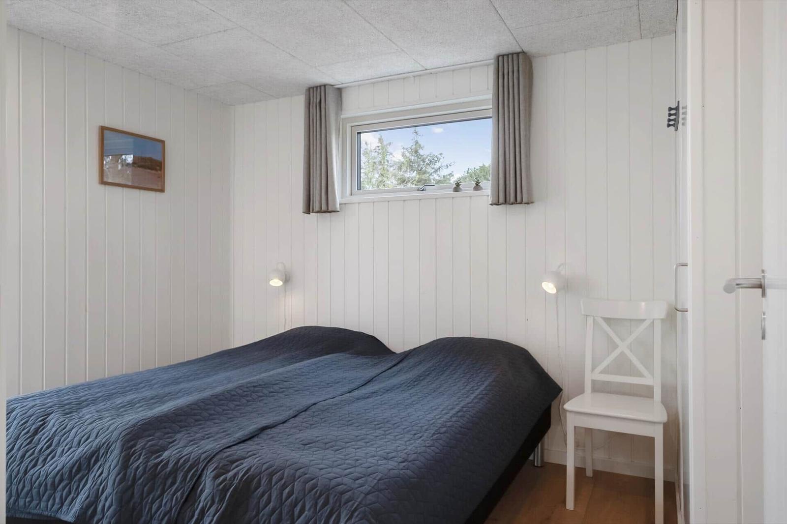 Schlafzimmer mit Bett, Fenster und Stuhl. Wände weiß verkleidet, Decke mit Paneelen.
