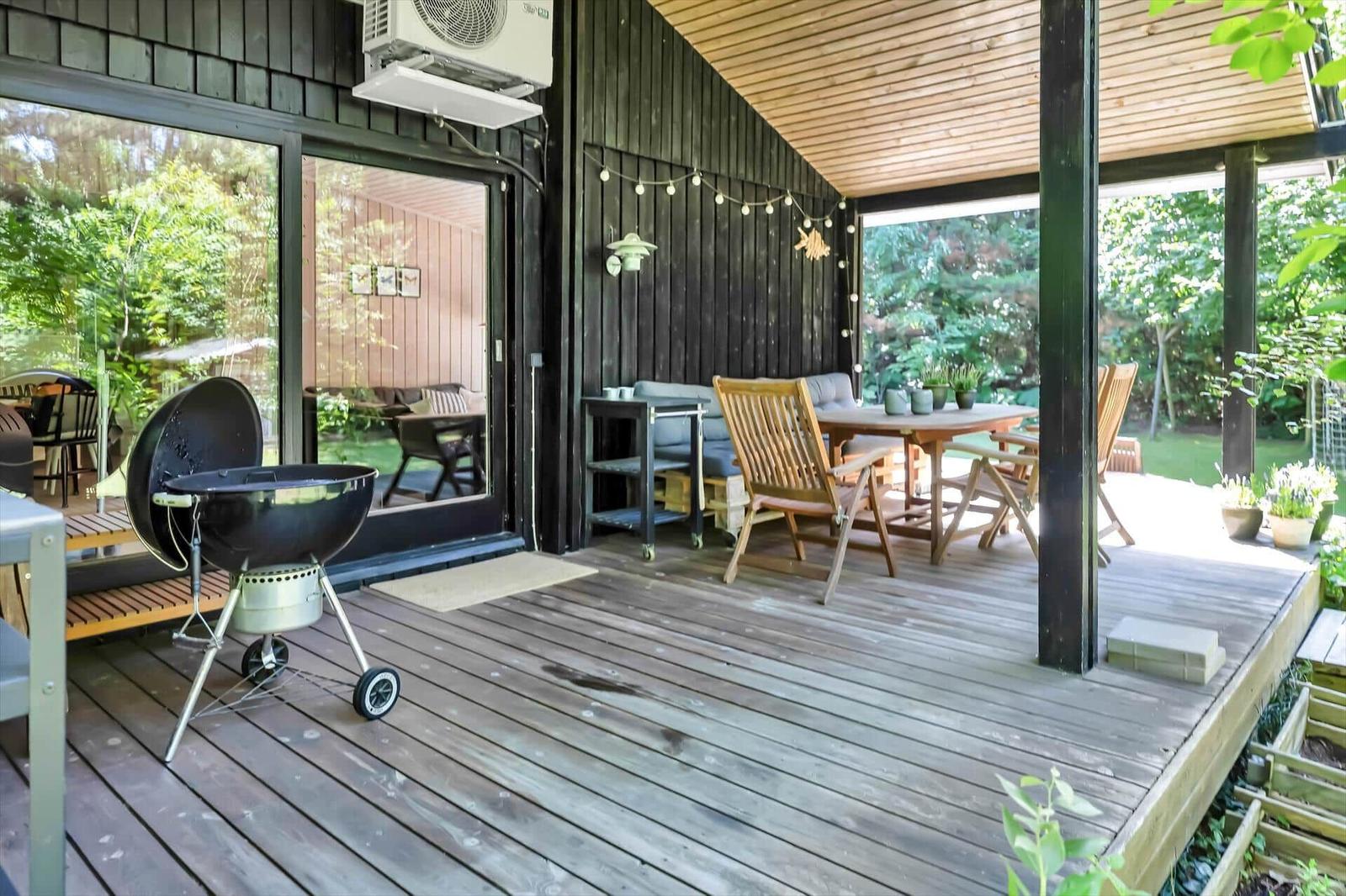 Terrasse mit Grill, Tisch und Stühlen. Holzdecke und Pflanzen im Hintergrund.