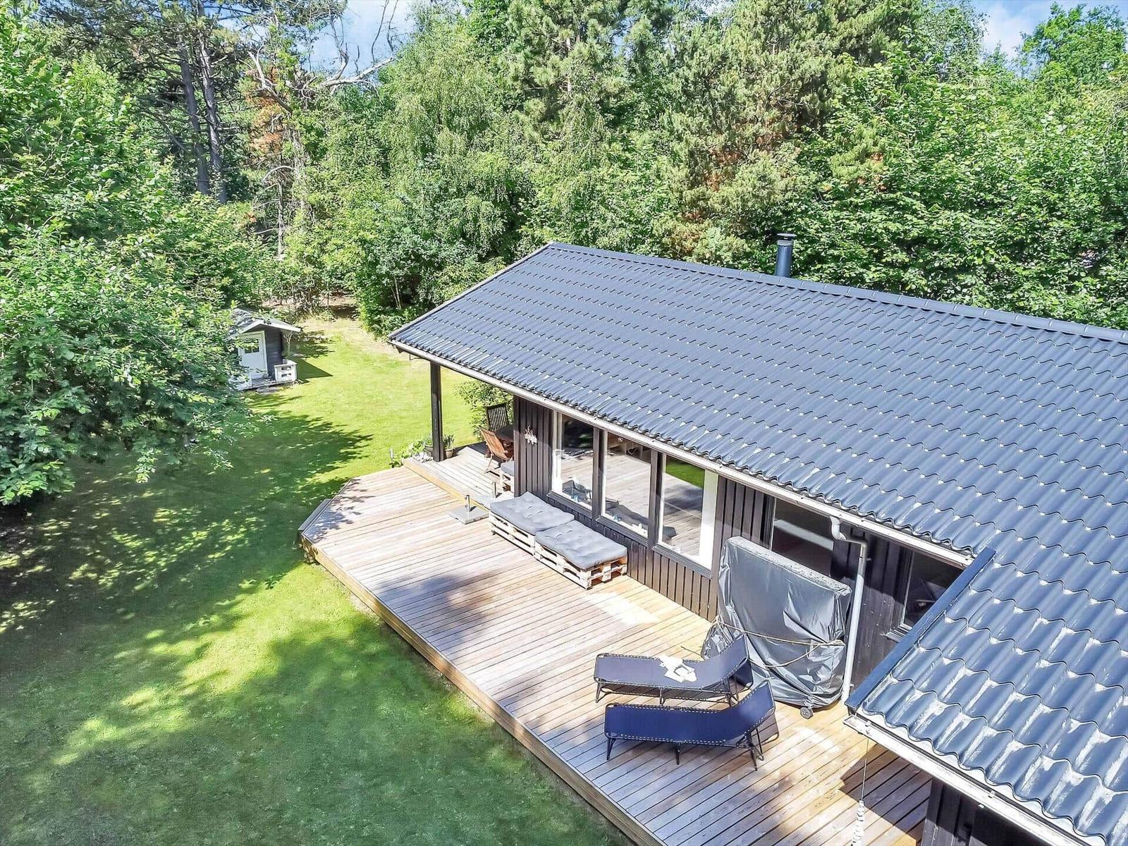 Holzterrasse mit Liegen und Grill, umgeben von grünem Rasen und Bäumen.
