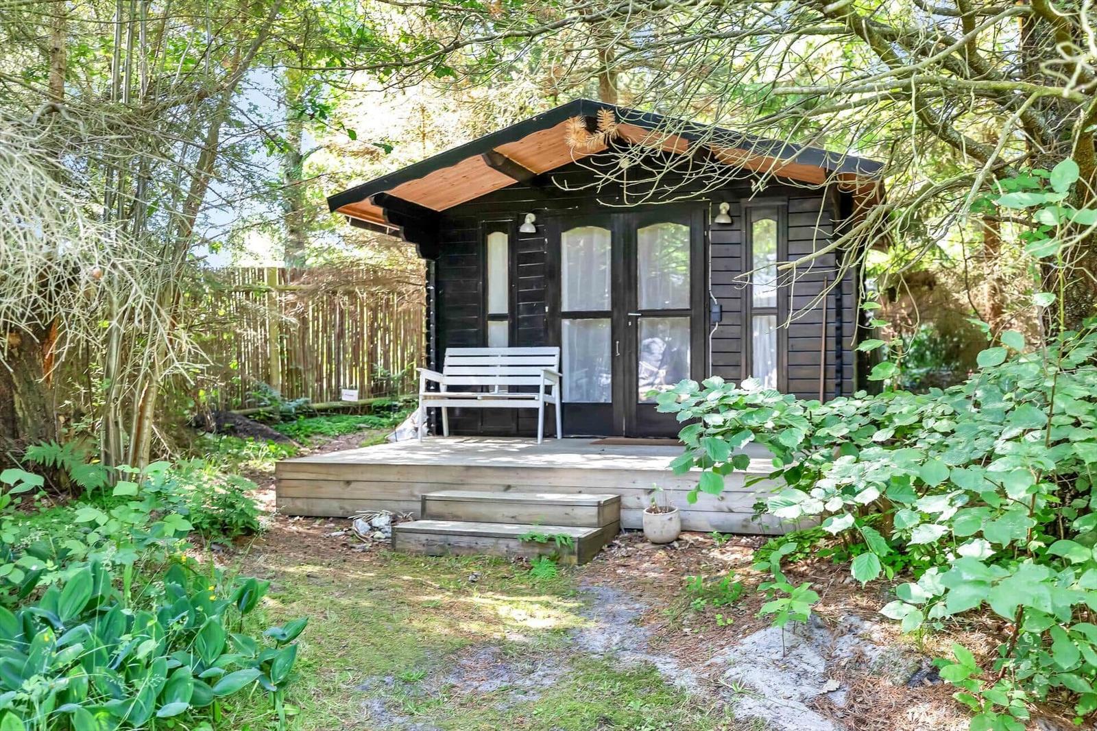 Holzhütte mit Terrasse und weißem Stuhl im Wald. Fenster mit weißen Vorhängen.