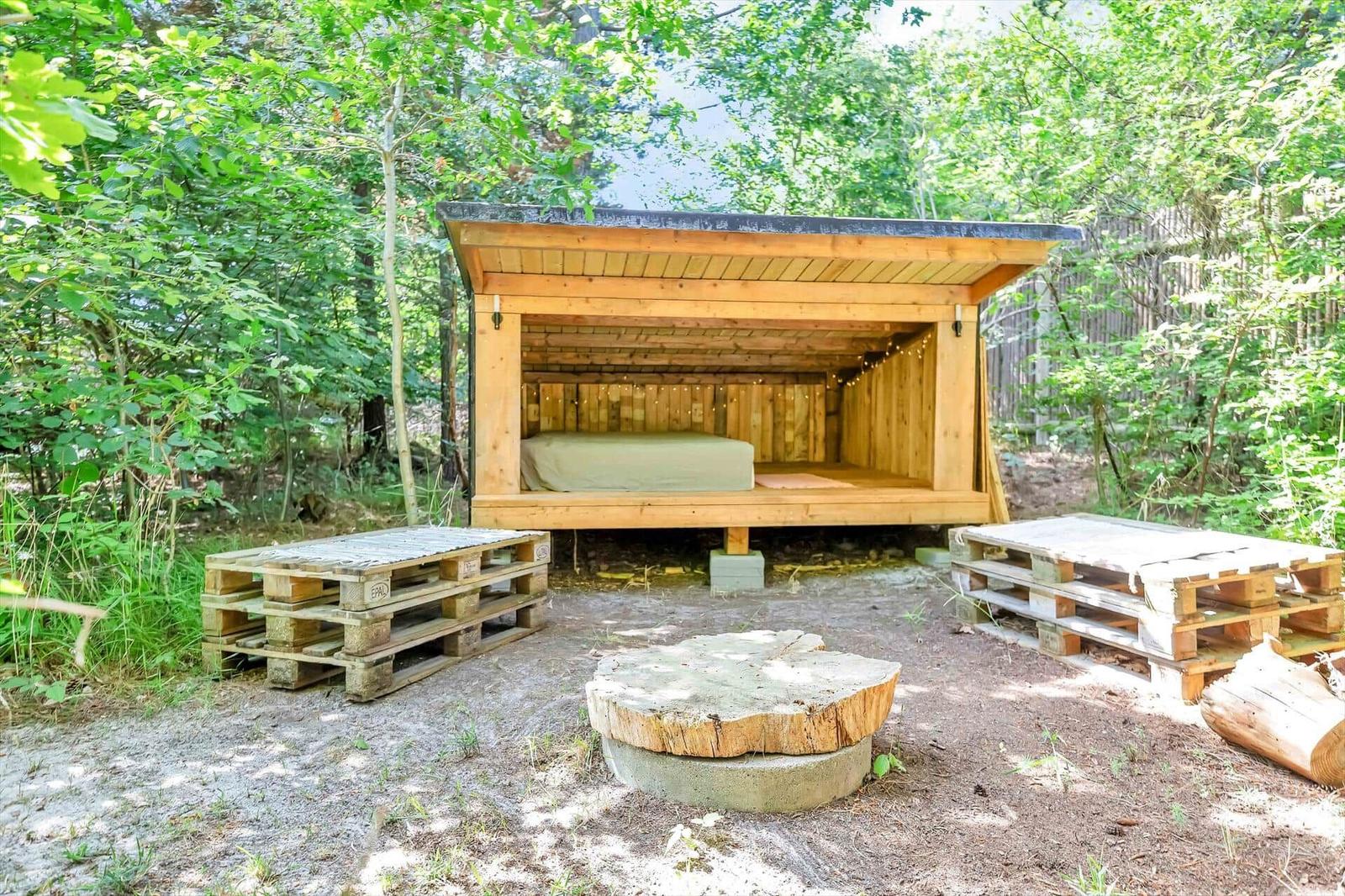 Holzkonstruktion mit Bett im Freien, umgeben von Bäumen und Paletten.