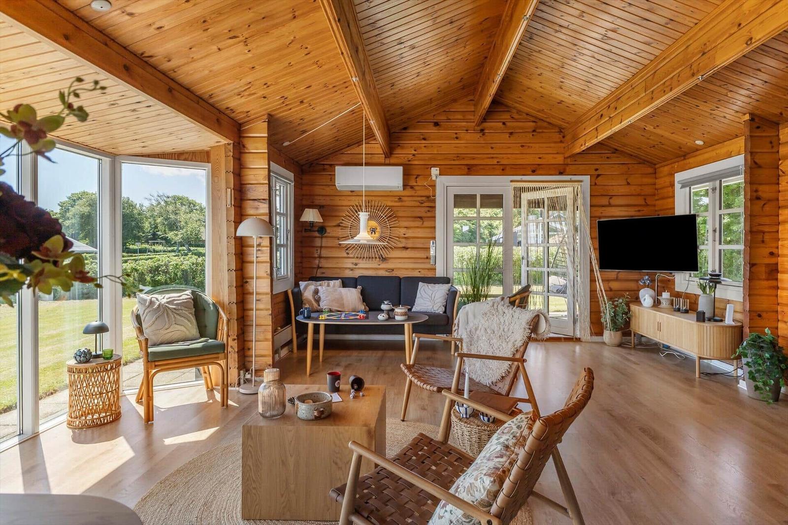 Wohnraum mit Holzdecke, Sofa, Stühlen und TV. Große Fenster bieten Blick auf Garten.