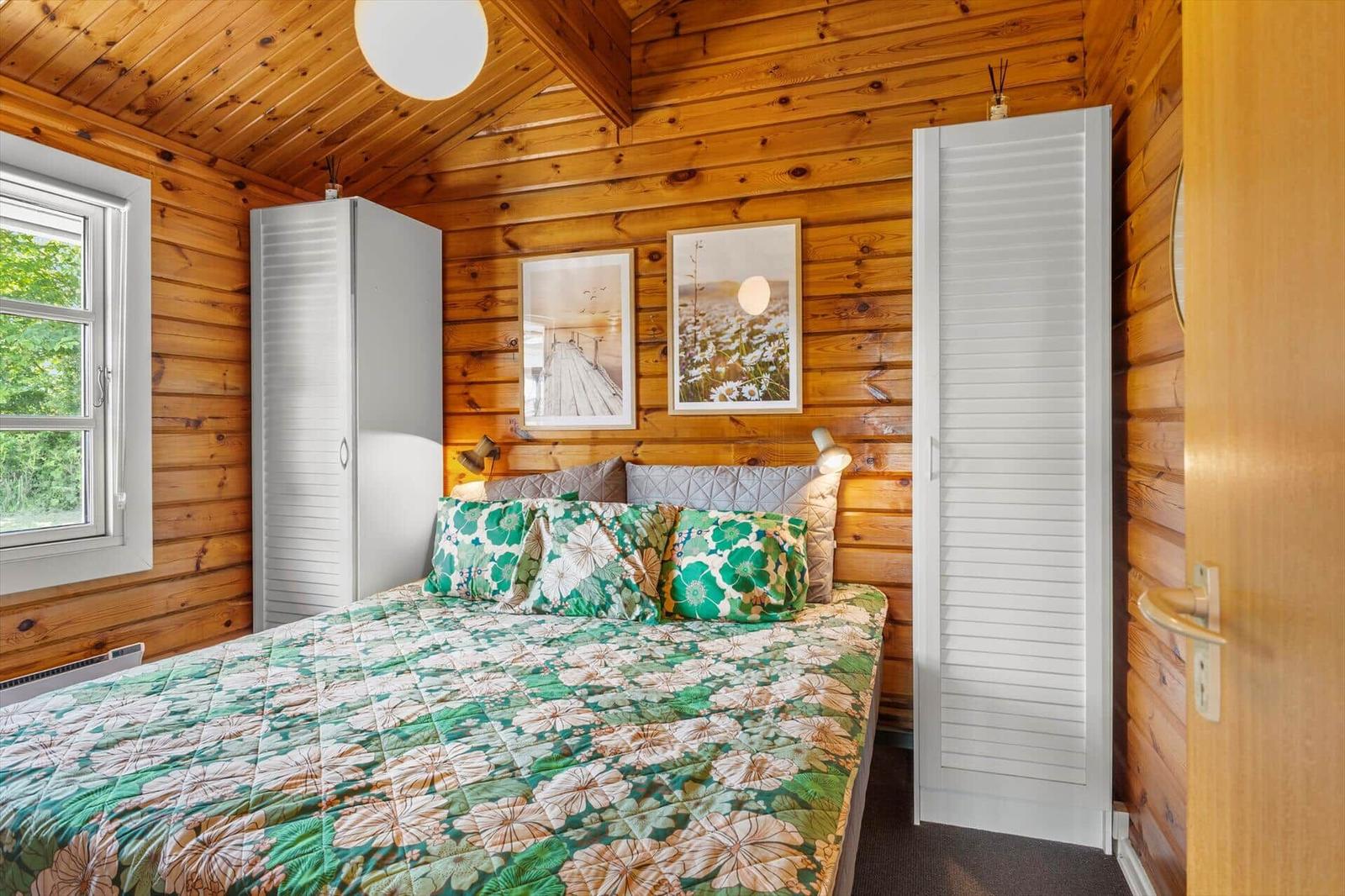 Schlafzimmer mit Holzwand, Bett mit floraler Bettwäsche, Fenster und weiße Schranktüren.